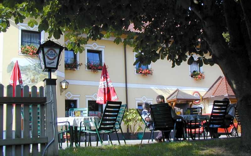 Ein Biergarten mit Tischen und Stühlen unter einem Baum vor einem gelben Gebäude mit Blumenkästen.