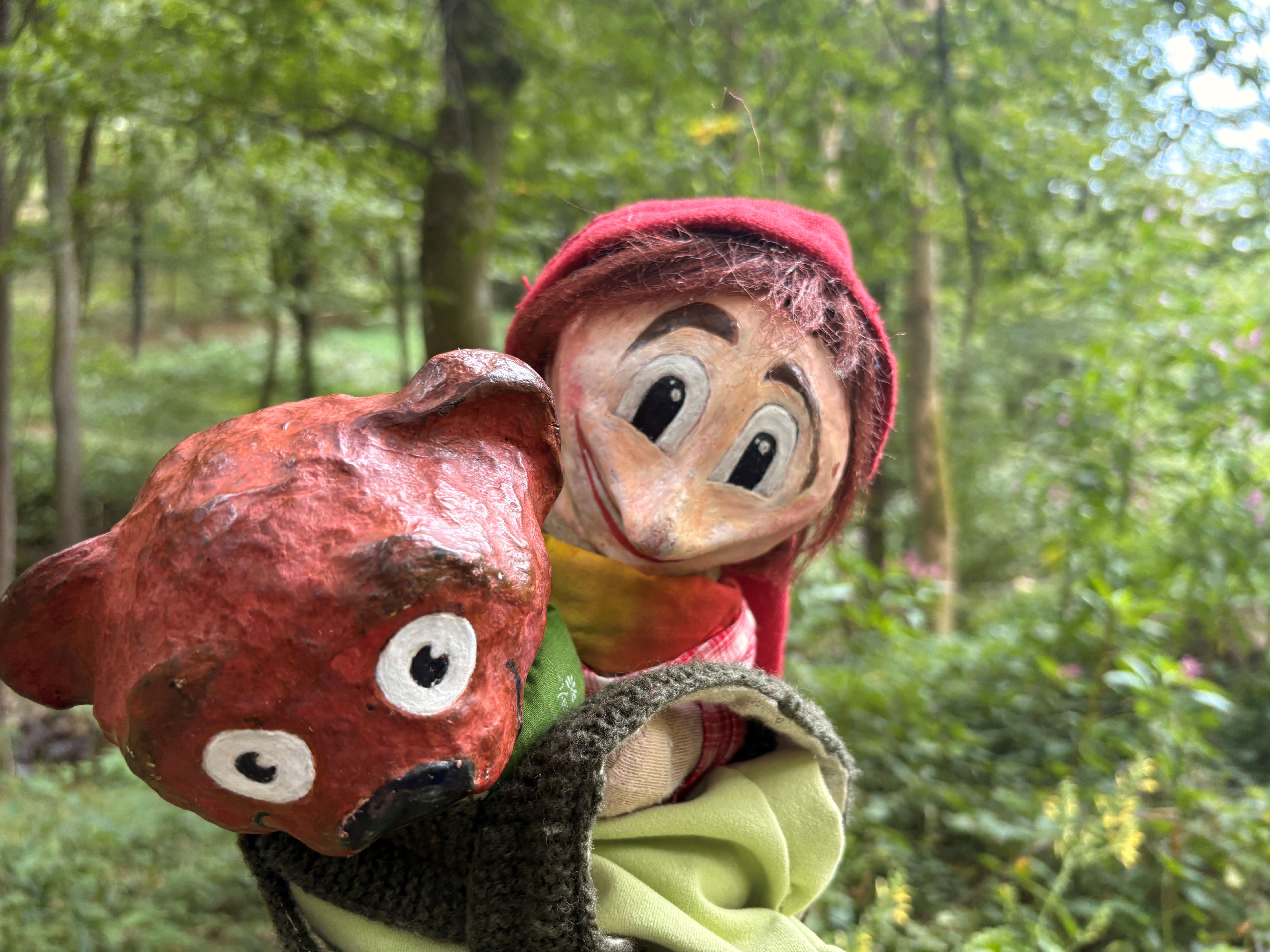 Eine Handpuppe mit rotem Hut und einem roten Bären in einem Wald.