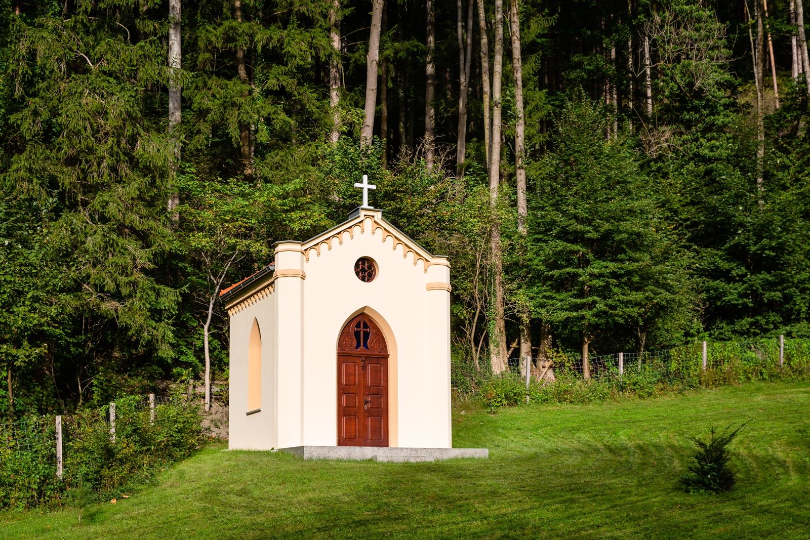 Eine kleine Kapelle mit einem Kreuz auf dem Dach, umgeben von Bäumen und Gras.