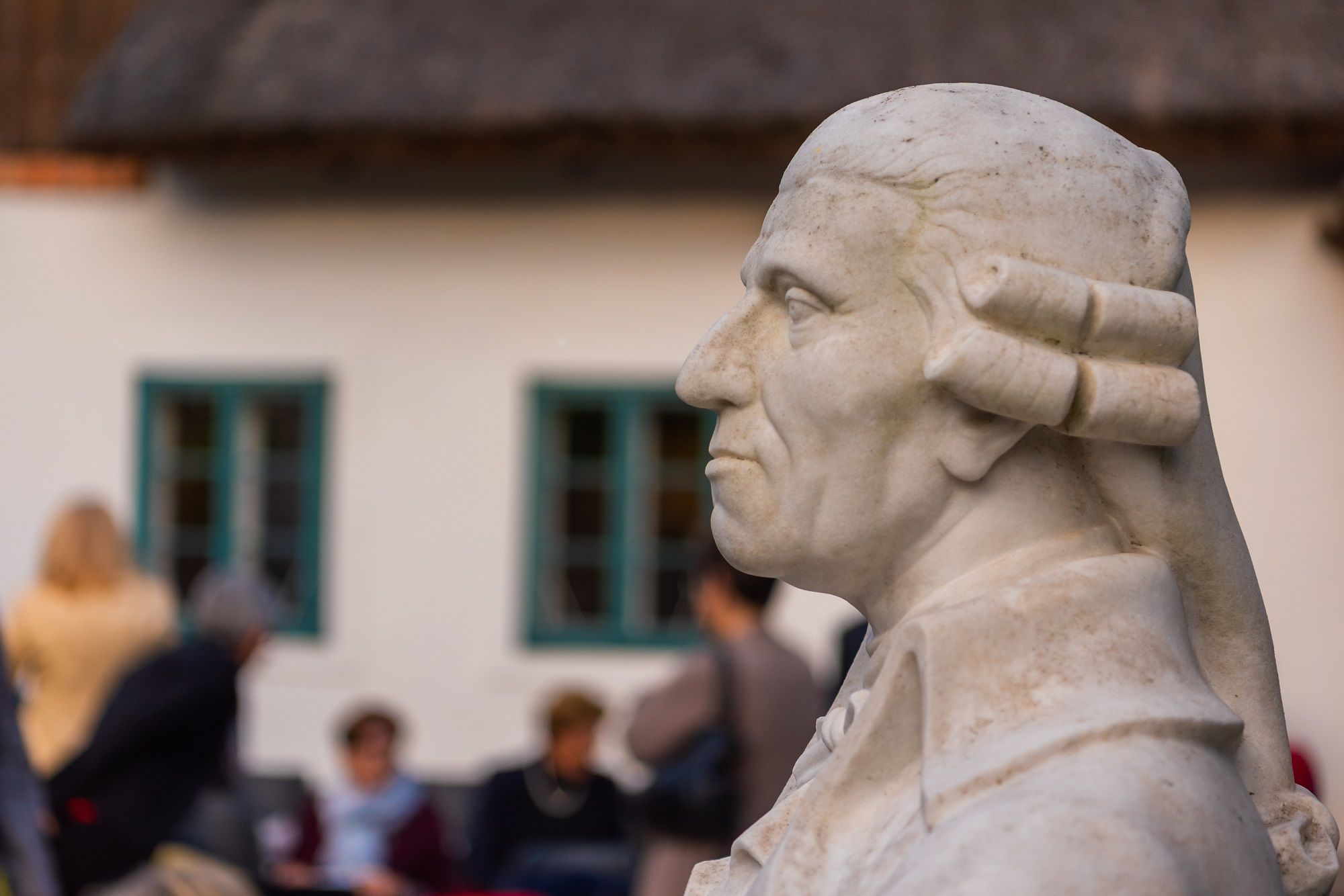 Büste eines Mannes im Profil vor einem Gebäude mit Menschen im Hintergrund.