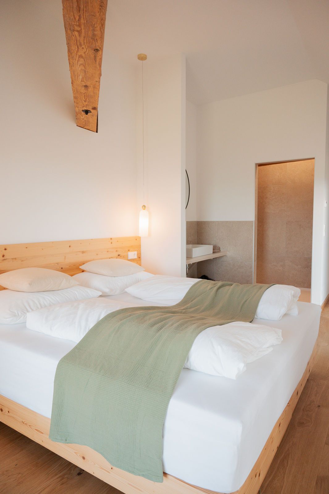 Ein modernes Schlafzimmer mit einem großen Bett, weißen Bettlaken und einem grünen Überwurf. Eine Holzdecke und eine dezente Beleuchtung sind sichtbar.