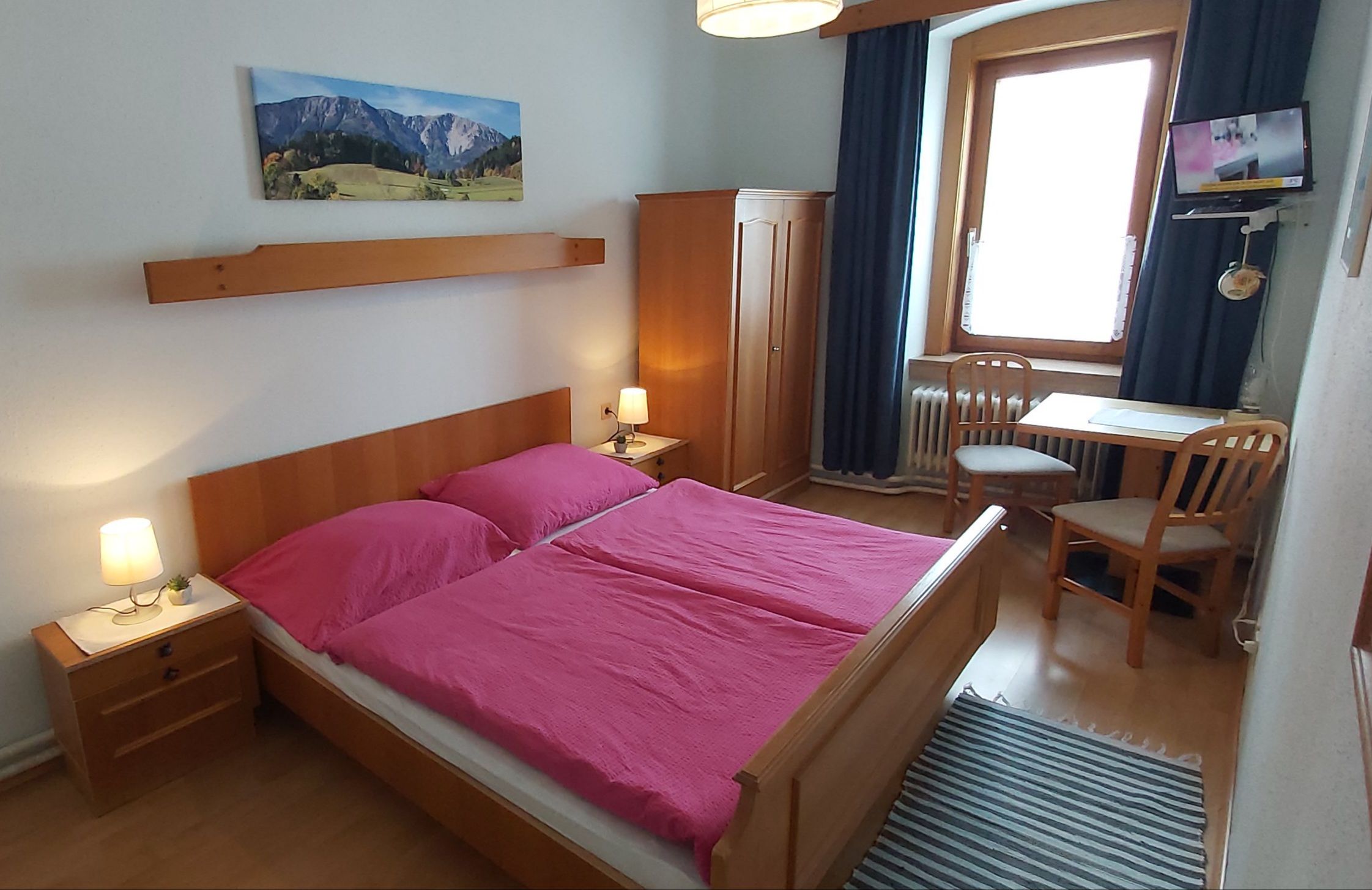 Ein gemütliches Doppelzimmer mit Holzmöbeln, einem Doppelbett mit pinker Bettwäsche, einem Tisch mit zwei Stühlen und einem kleinen Fernseher.