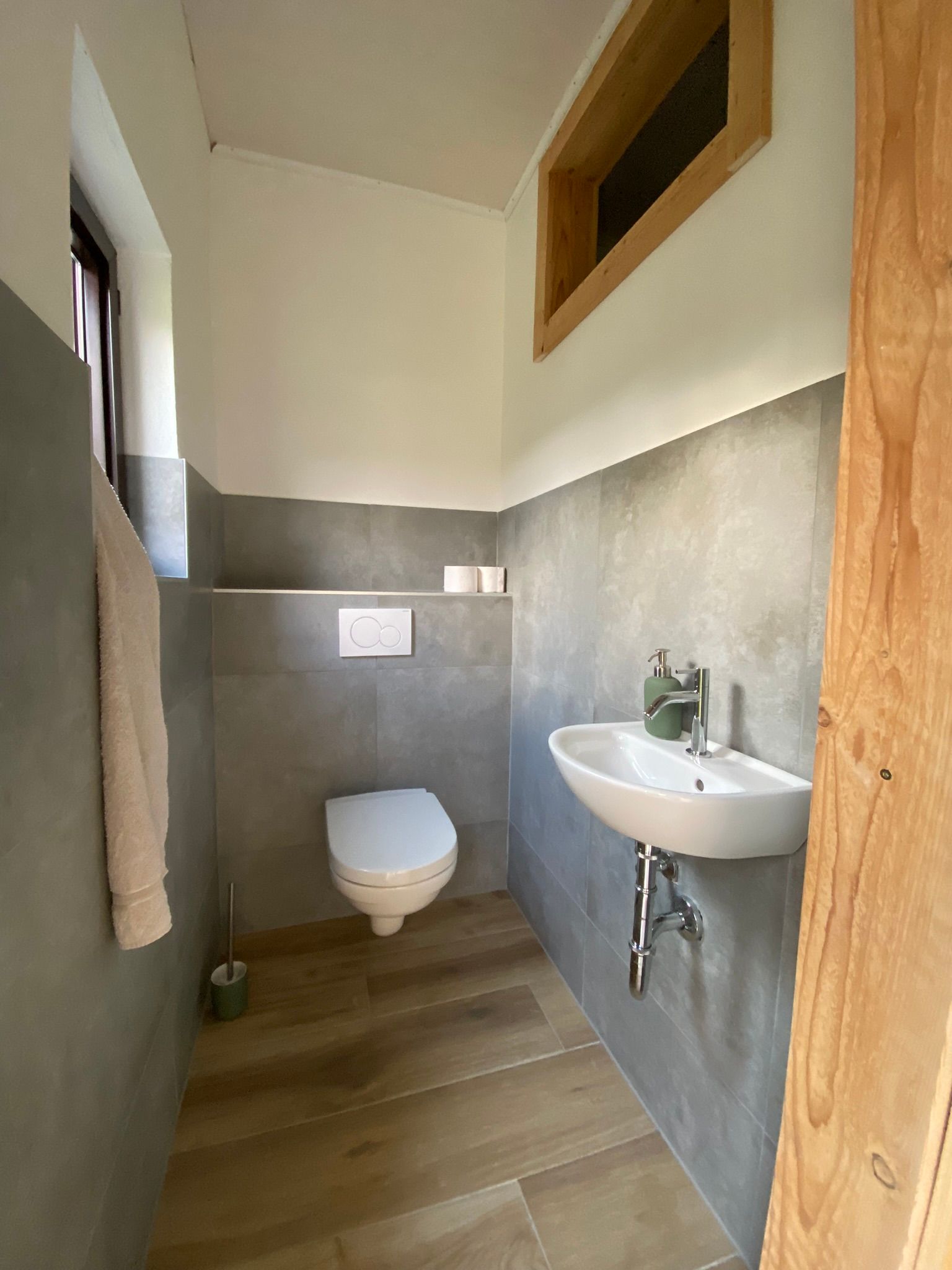 WC mit Wandtoilette, kleinem Waschbecken und grauen Fliesen.