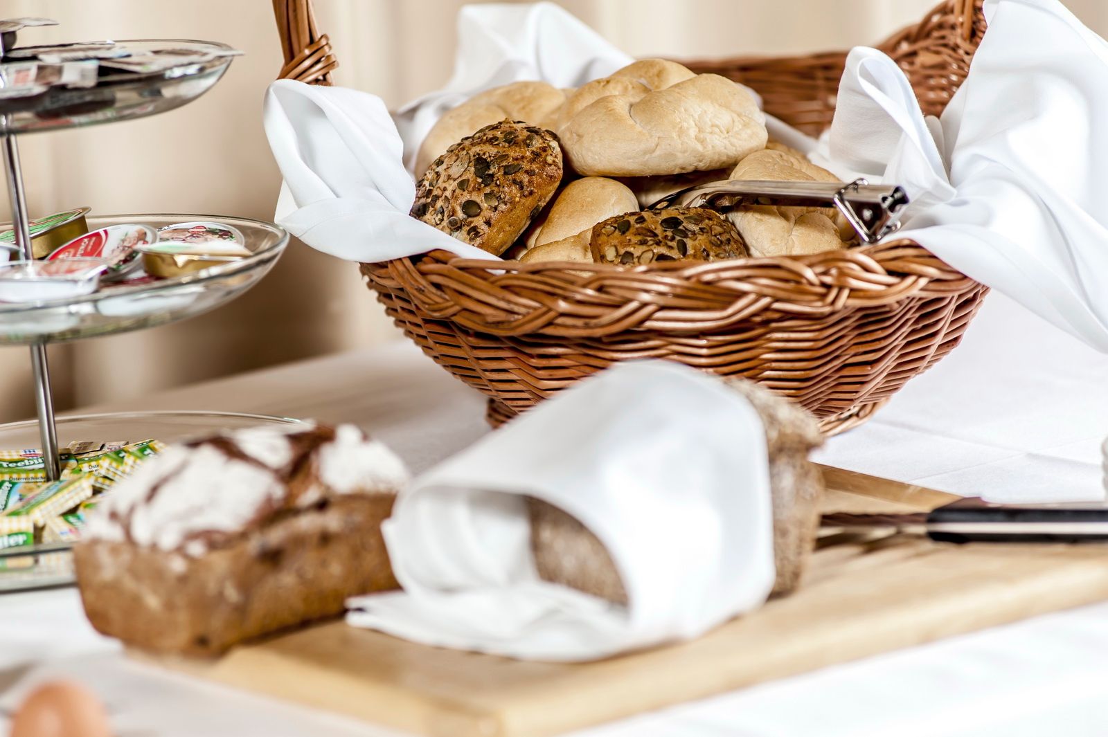 Ein Frühstückstisch mit einem Korb voller Brötchen, einem Brotschneidebrett und einem Tablett mit Marmeladen.