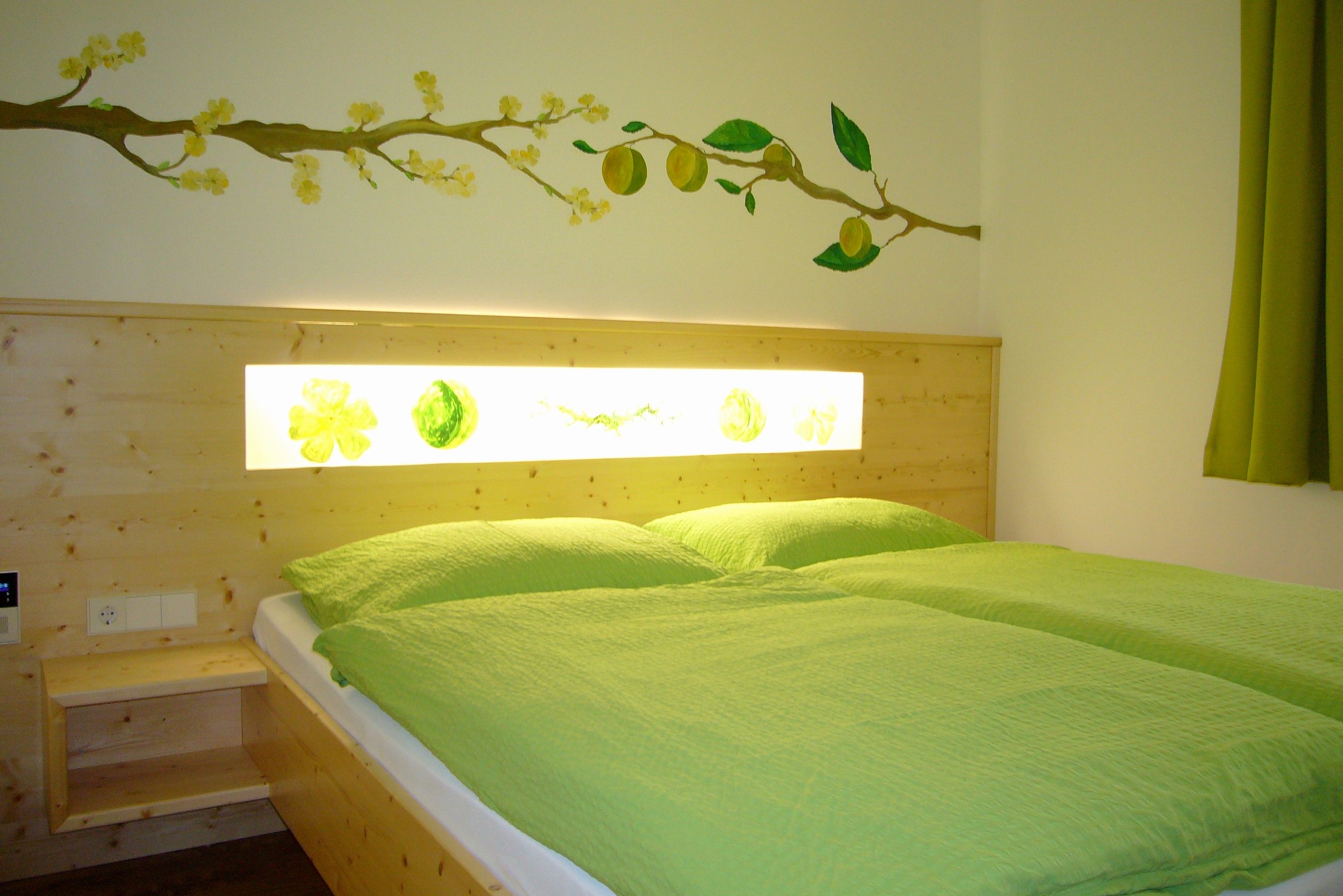 Ein gemütliches Zimmer mit einem Doppelbett, grüner Bettwäsche und einer Wanddekoration mit gelben Blüten und Früchten.