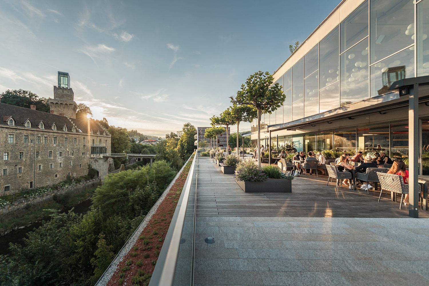Panorama einer Flusslandschaft mit Schloss und moderner Architektur, umgeben von grünen Hügeln.