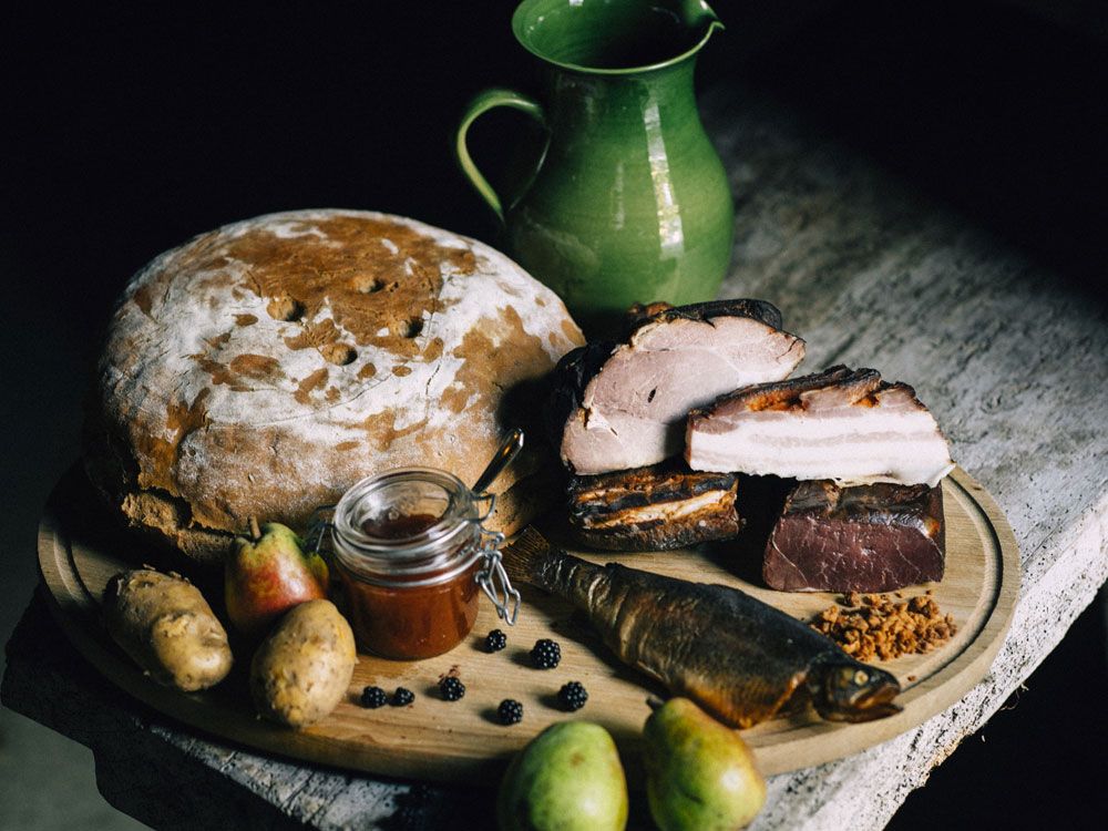 Ein rustikales Arrangement mit Brot, Fleisch, Fisch, Birnen, Kartoffeln und Marmelade auf einem Holztisch.