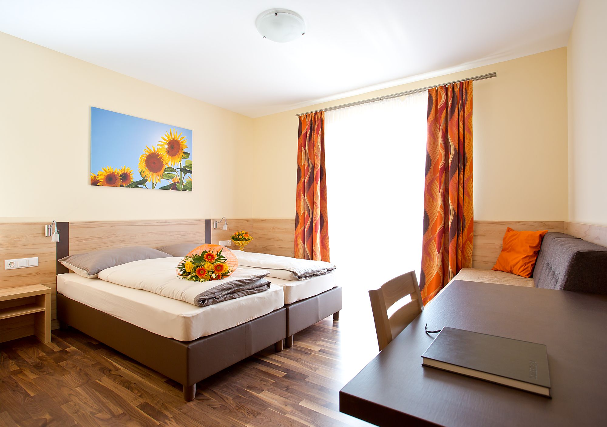 Ein helles Gästezimmer mit zwei Betten, einem Tisch, einem Sofa und einem Bild von Sonnenblumen an der Wand.