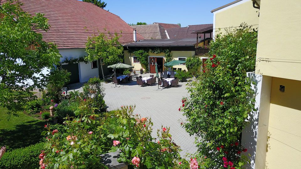 Innenhof eines Winzerhofs mit Tischen, Stühlen und Sonnenschirmen, umgeben von blühenden Pflanzen und Gebäuden mit roten Dächern.