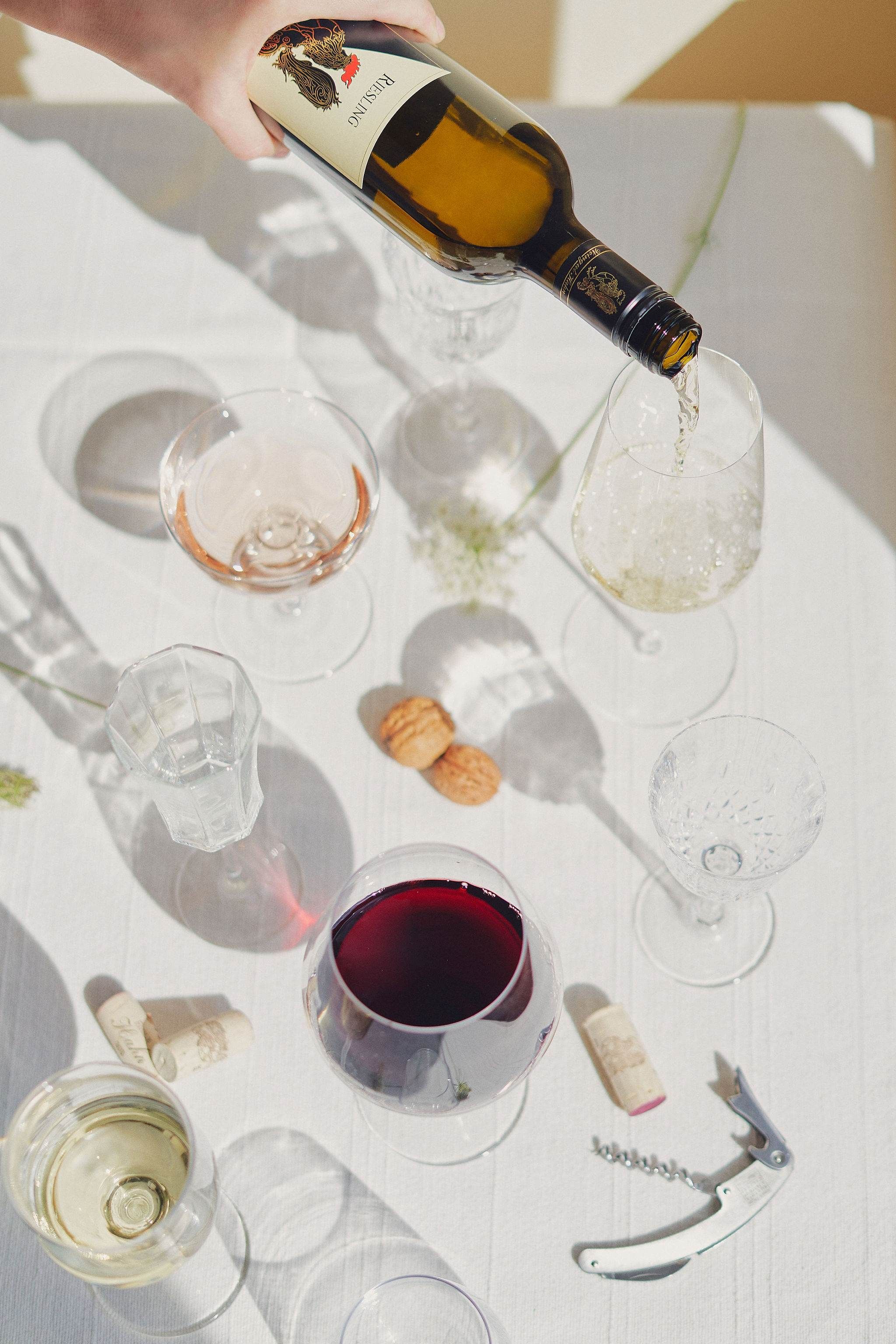 Weinflasche wird in ein Glas auf einem Tisch mit verschiedenen Weingläsern und Korken gegossen.