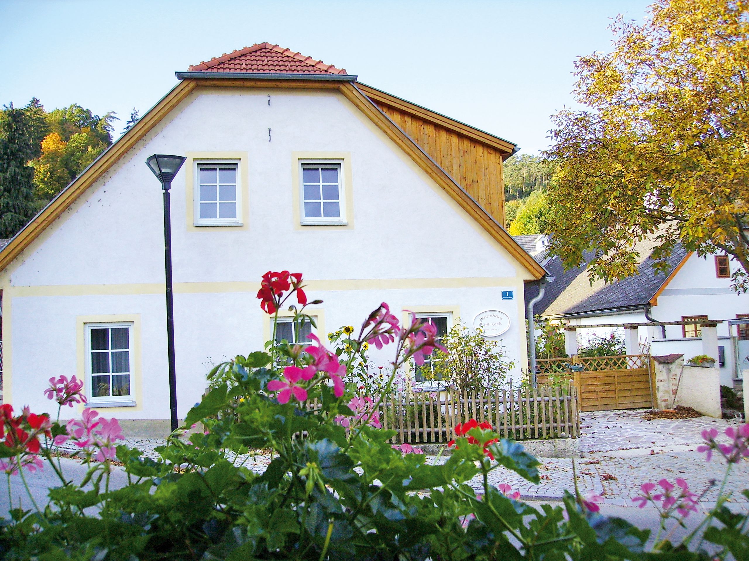 Ein weißes Ferienhaus mit roten Dachziegeln und einem Gartenzaun, umgeben von blühenden Blumen und Bäumen im Herbst.