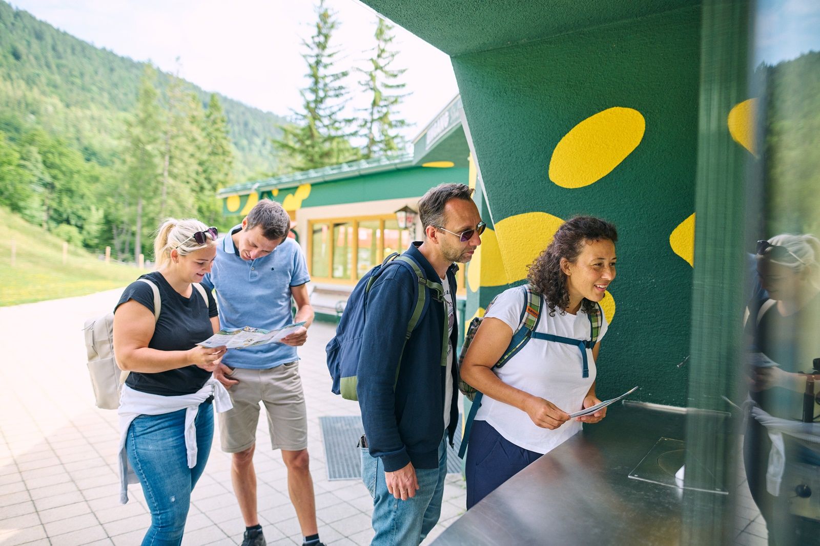 Vier Personen stehen an einem Ticketschalter im Freien, umgeben von grüner Landschaft.