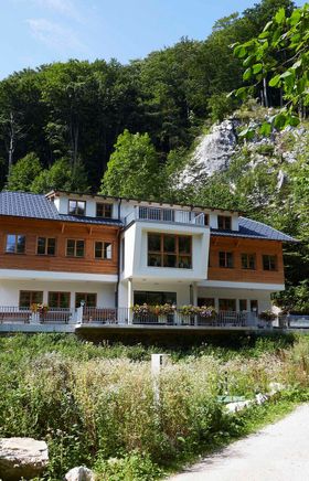 Hütte liegt zwischen vielen Bäumen am Beginn einer Felsschlucht, im Vordergrund ist ein Weg.