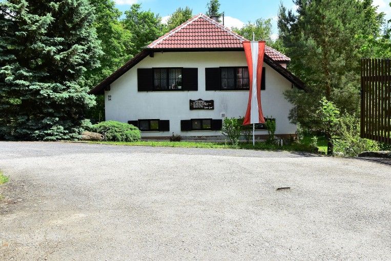 Ein weißes Haus mit rotem Dach und österreichischer Flagge davor, umgeben von Bäumen.