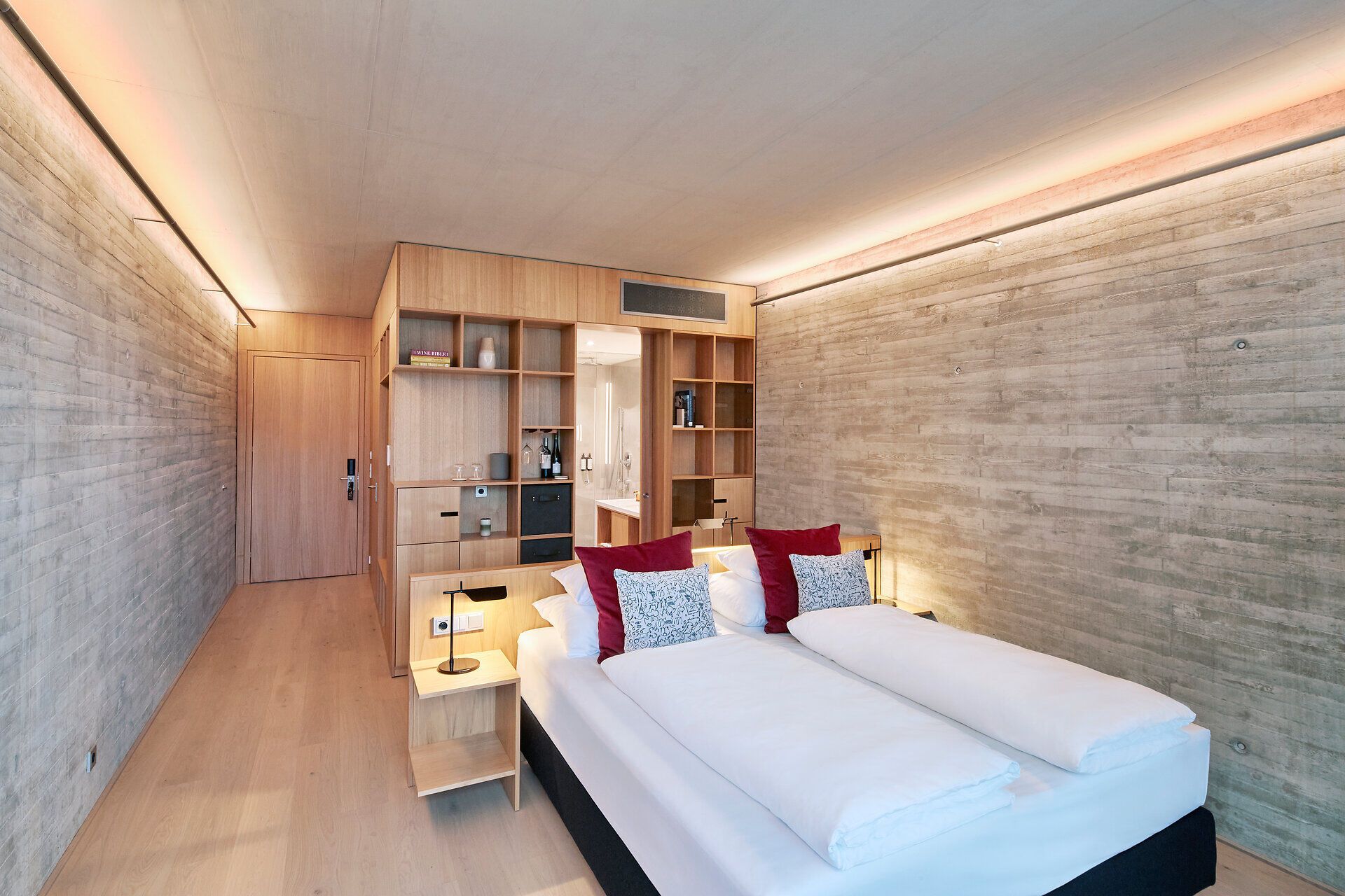 Ein hochmodernes Hotelzimmer in minimalistischem Design. Die linke Raumhälfte besteht aus hellem Naturholz, das nahtlos vom Boden in eine deckenhohe Regalwand und die Zimmertür übergeht. Rechts bildet eine Wand in Sichtbeton-Optik einen kühlen Kontrast. Das Doppelbett im Vordergrund ist schlicht weiß bezogen und wird durch zwei weinrote sowie zwei gemusterte Zierkissen akzentuiert. Eine dezente, indirekte Beleuchtung an der Decke sorgt für eine warme Atmosphäre.