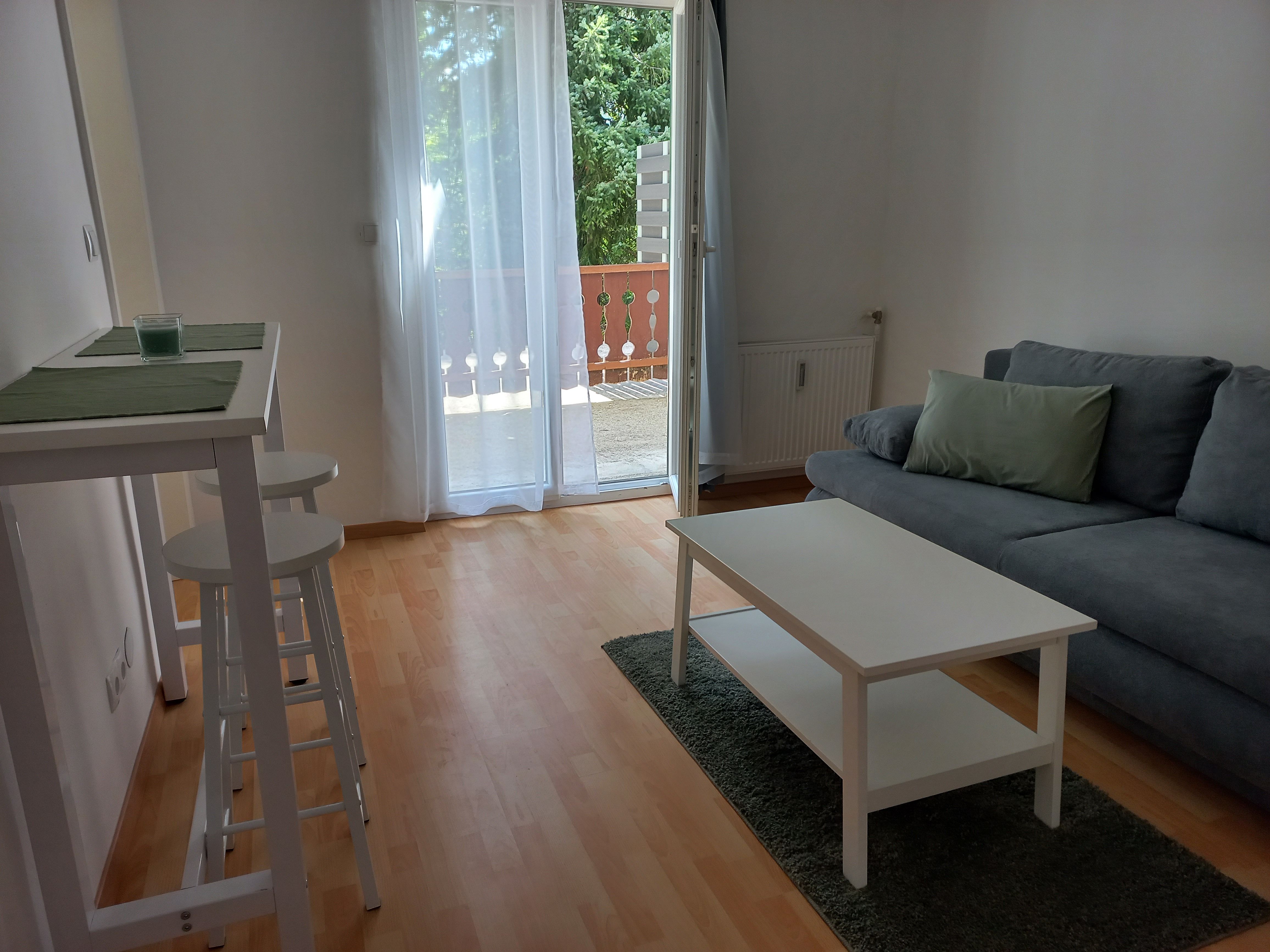 Modernes Wohnzimmer mit grauem Sofa, weißem Couchtisch und Barhockern. Balkon mit Glas-Schiebetür im Hintergrund.