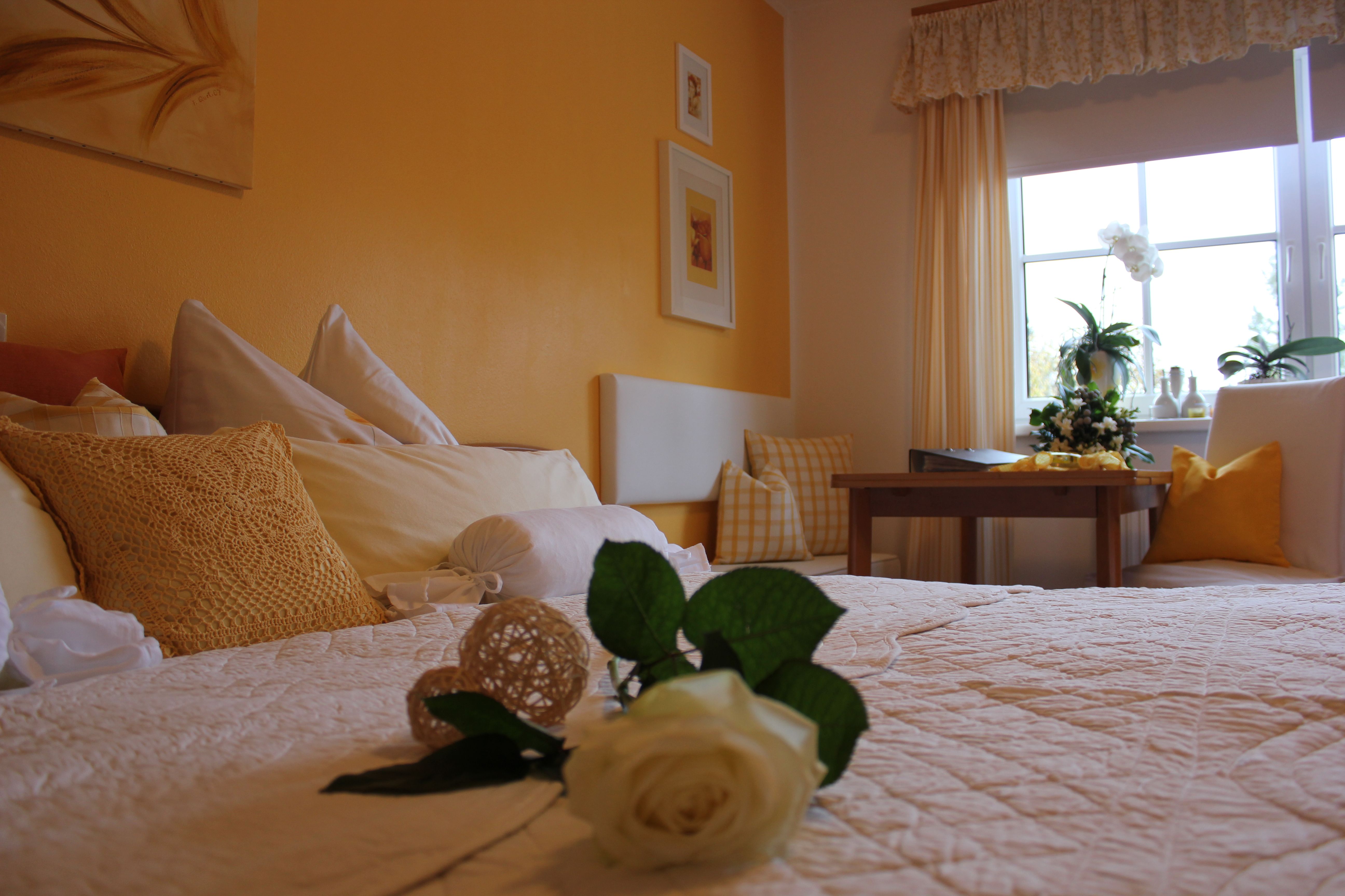 Gemütliches Schlafzimmer mit gelben Wänden, einem Bett mit weißen und gelben Kissen, einem Tisch und einem Fenster mit Pflanzen.