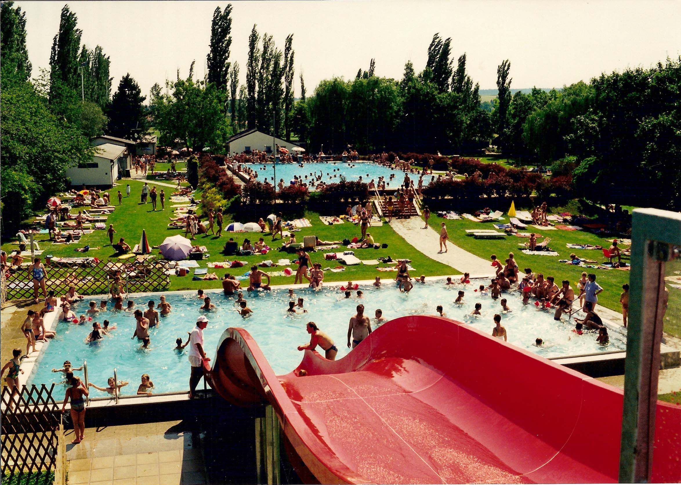Ein Freibad mit mehreren Pools, einer roten Wasserrutsche und vielen Menschen, die im Wasser schwimmen oder auf der Wiese liegen.