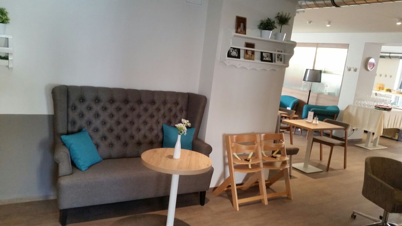 Innenansicht eines gemütlichen Cafés mit grauem Sofa, Holztischen und blauen Kissen.