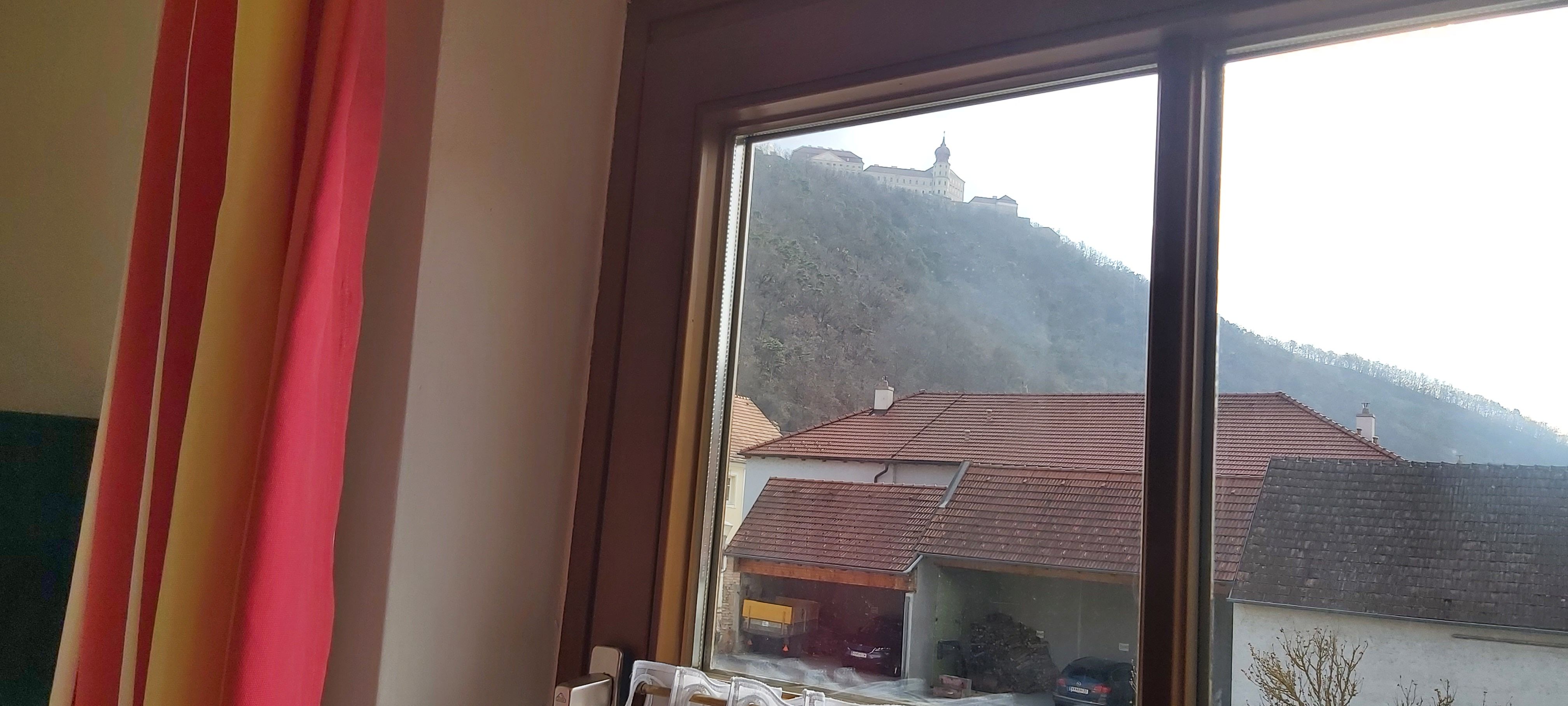 Blick aus einem Fenster auf ein Gebäude auf einem Hügel und Häuser mit roten Dächern im Vordergrund.