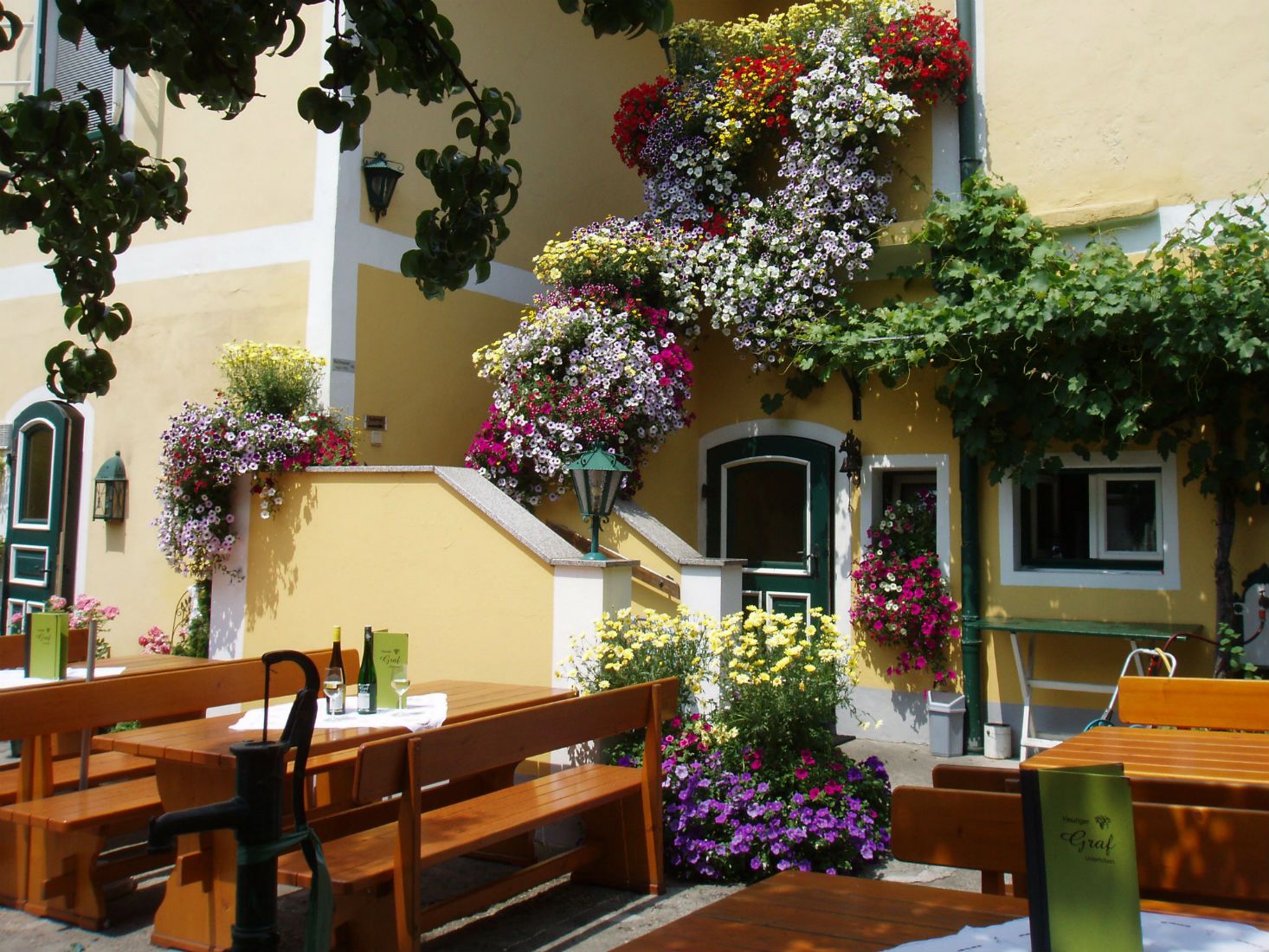 Ein gemütlicher Heurigengarten mit Holzbänken, Tischen und blühenden Blumen an einem gelben Gebäude.