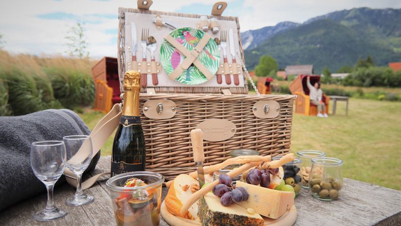 Picknickkorb mit Käse, Trauben und Sektflasche auf einem Tisch in einer ländlichen Umgebung mit Bergen im Hintergrund.
