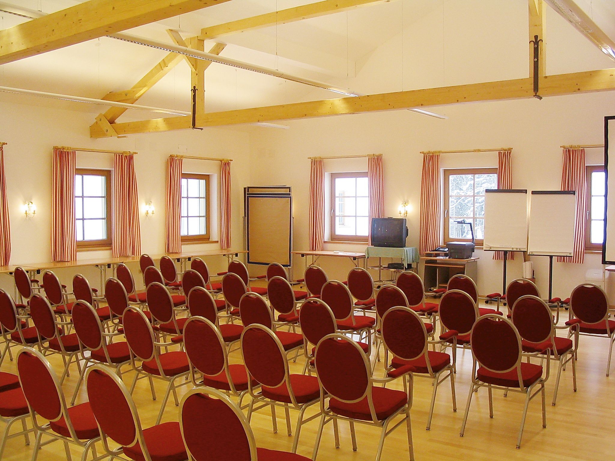Ein Konferenzraum mit roten Stühlen, Holzbalken und Fenstern mit gestreiften Vorhängen.