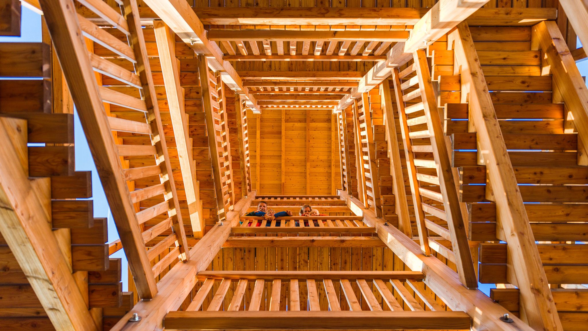 Blick von unten in einen hölzernen Aussichtsturm mit mehreren Ebenen und Treppen.