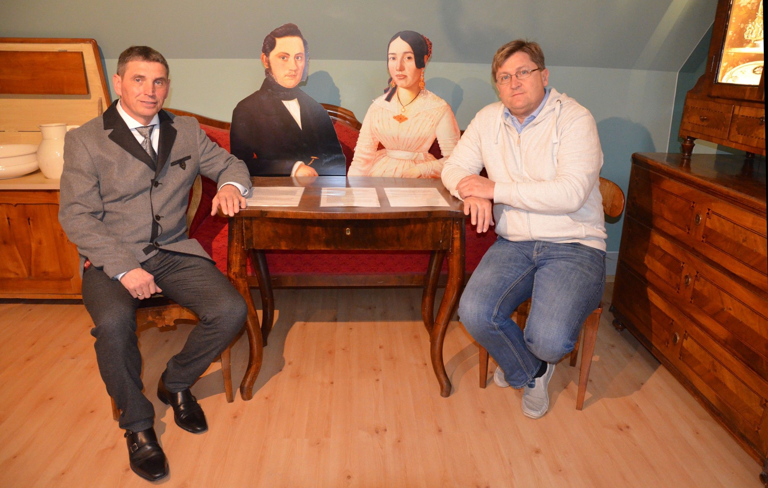 Zwei Männer sitzen an einem Tisch mit zwei Pappfiguren in einem Raum mit antiken Möbeln.