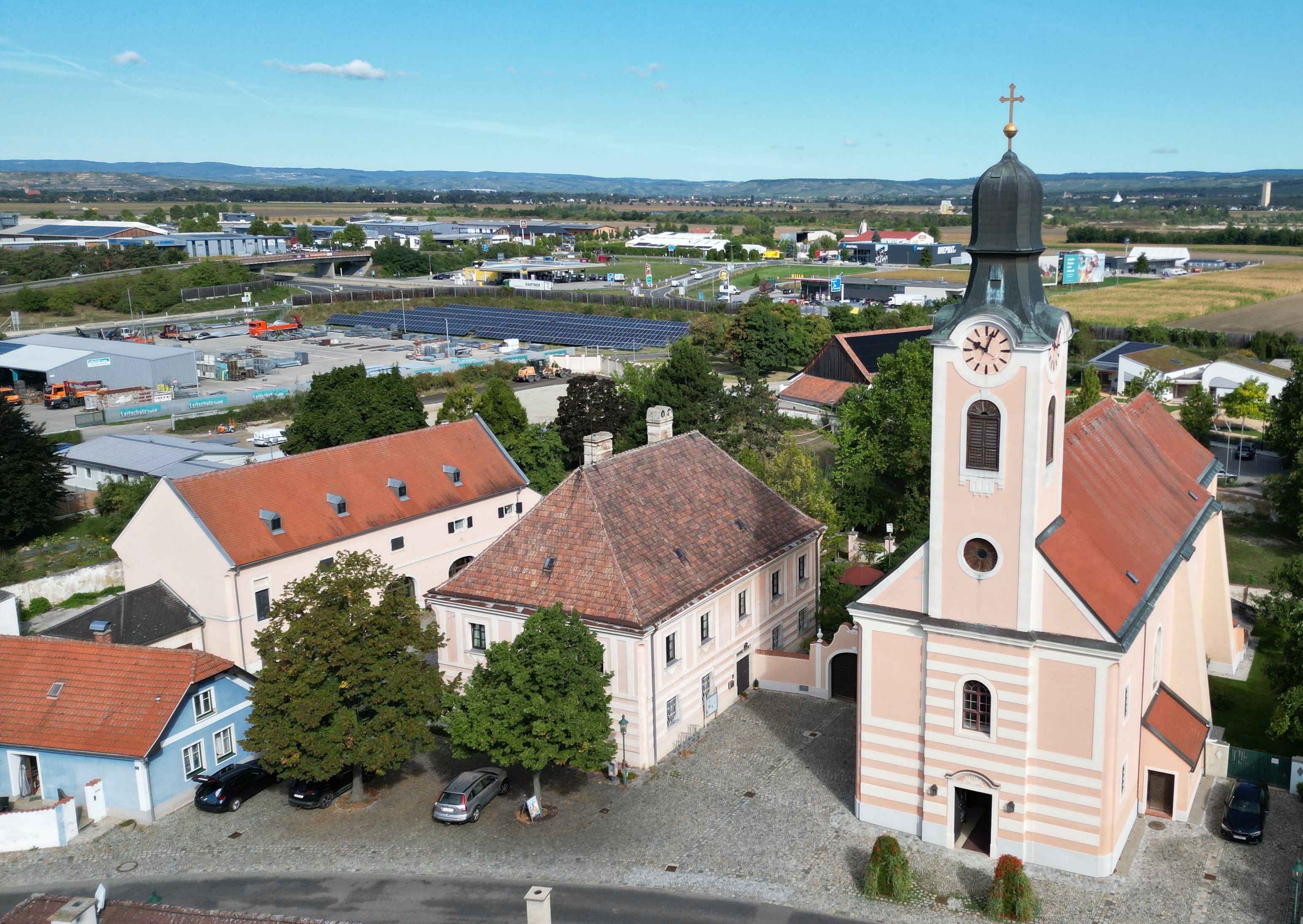 Luftaufnahme einer barocken Kirche mit Zwiebelturm und angrenzendem historischen Gebäude in Kirchberg am Wagram, mit Feldern und Gewerbegebiet im Hintergrund.