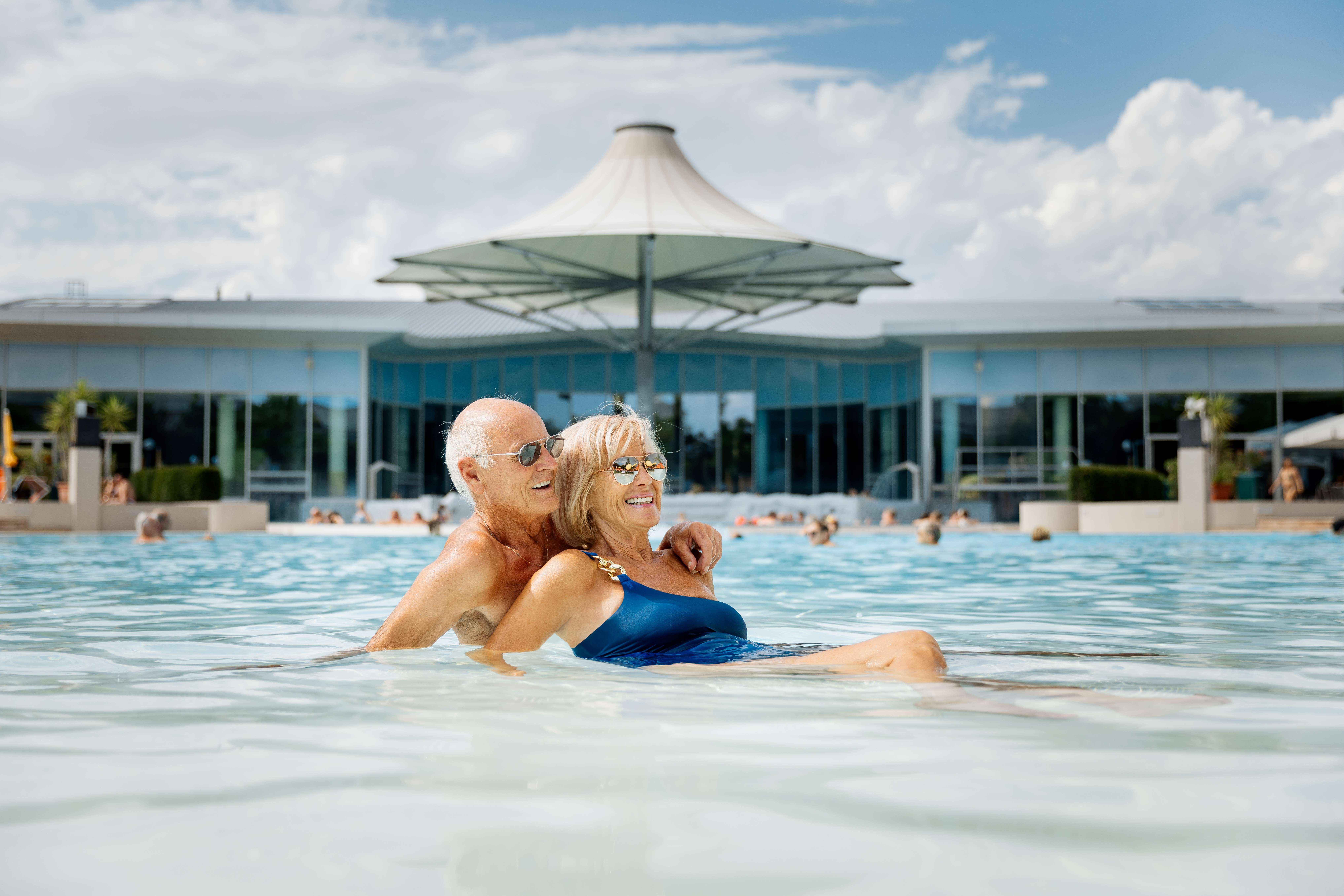 Ein älteres Paar entspannt im Außenpool eines modernen Thermenresorts bei sonnigem Wetter.
