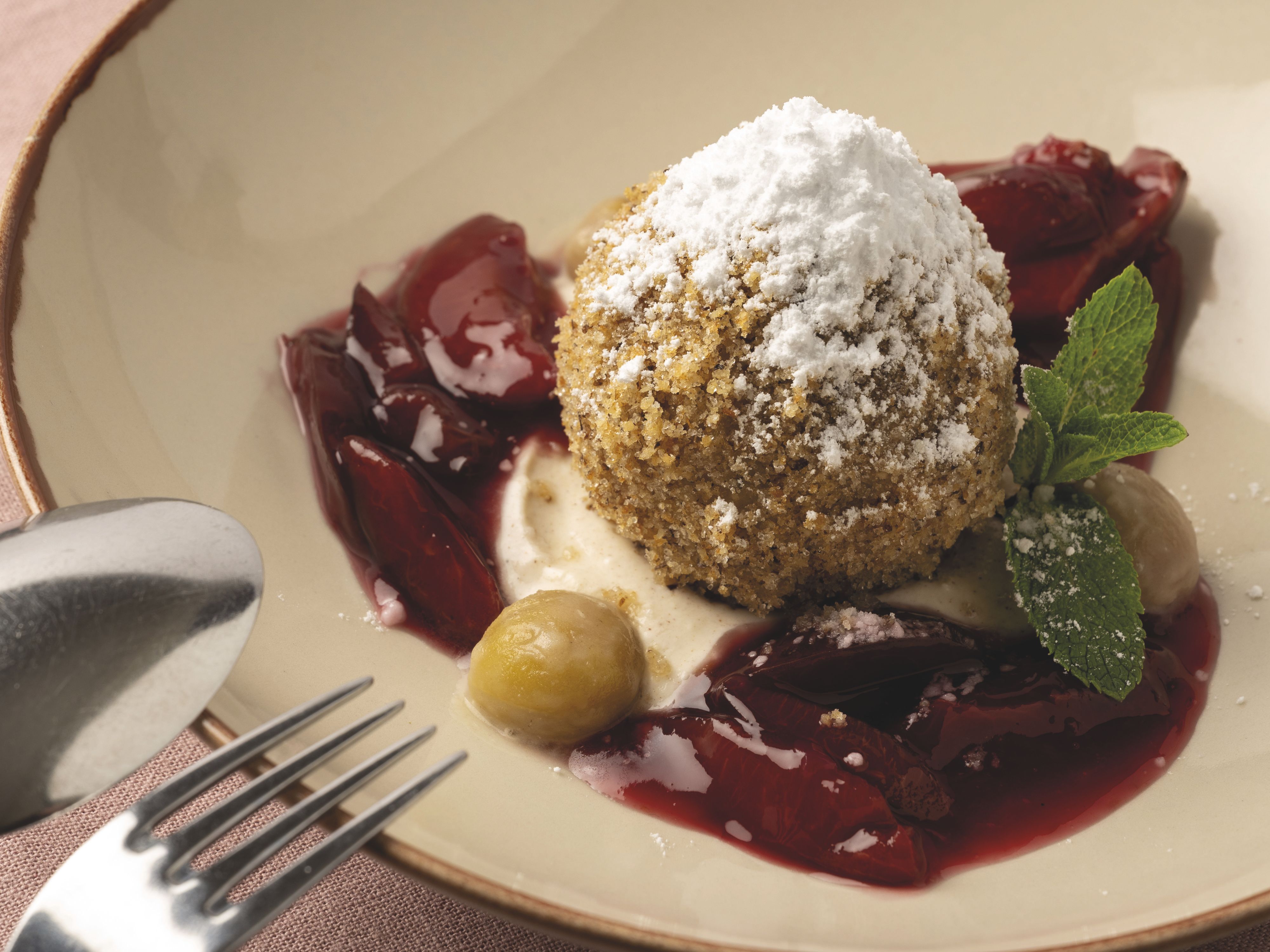 Ein Dessert mit einem Knödel, bestreut mit Puderzucker, auf einem Teller mit roten Früchten und Minzblättern dekoriert.