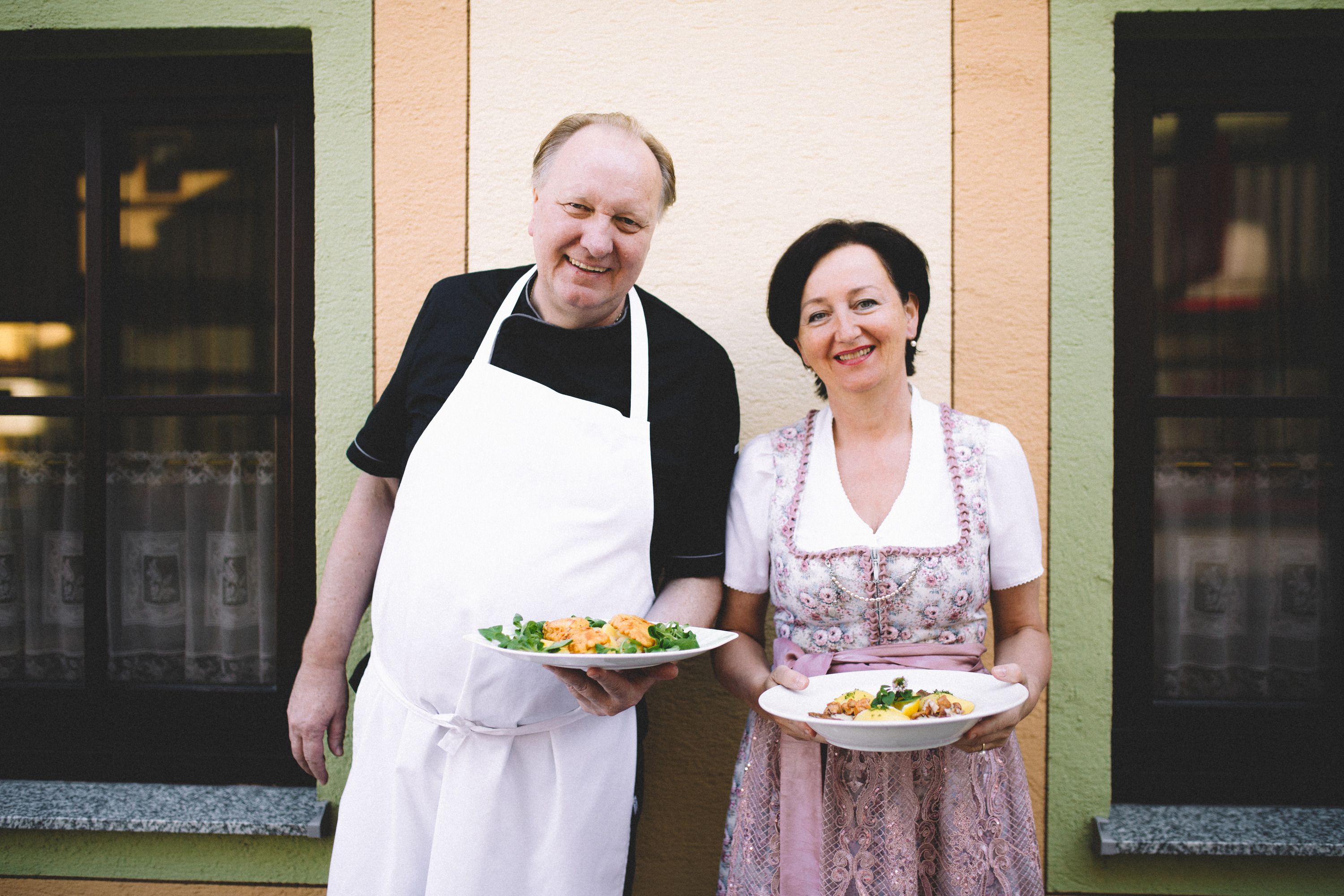 Ein Mann in Schürze und eine Frau in traditioneller Kleidung halten Teller mit Essen vor einem Gebäude.