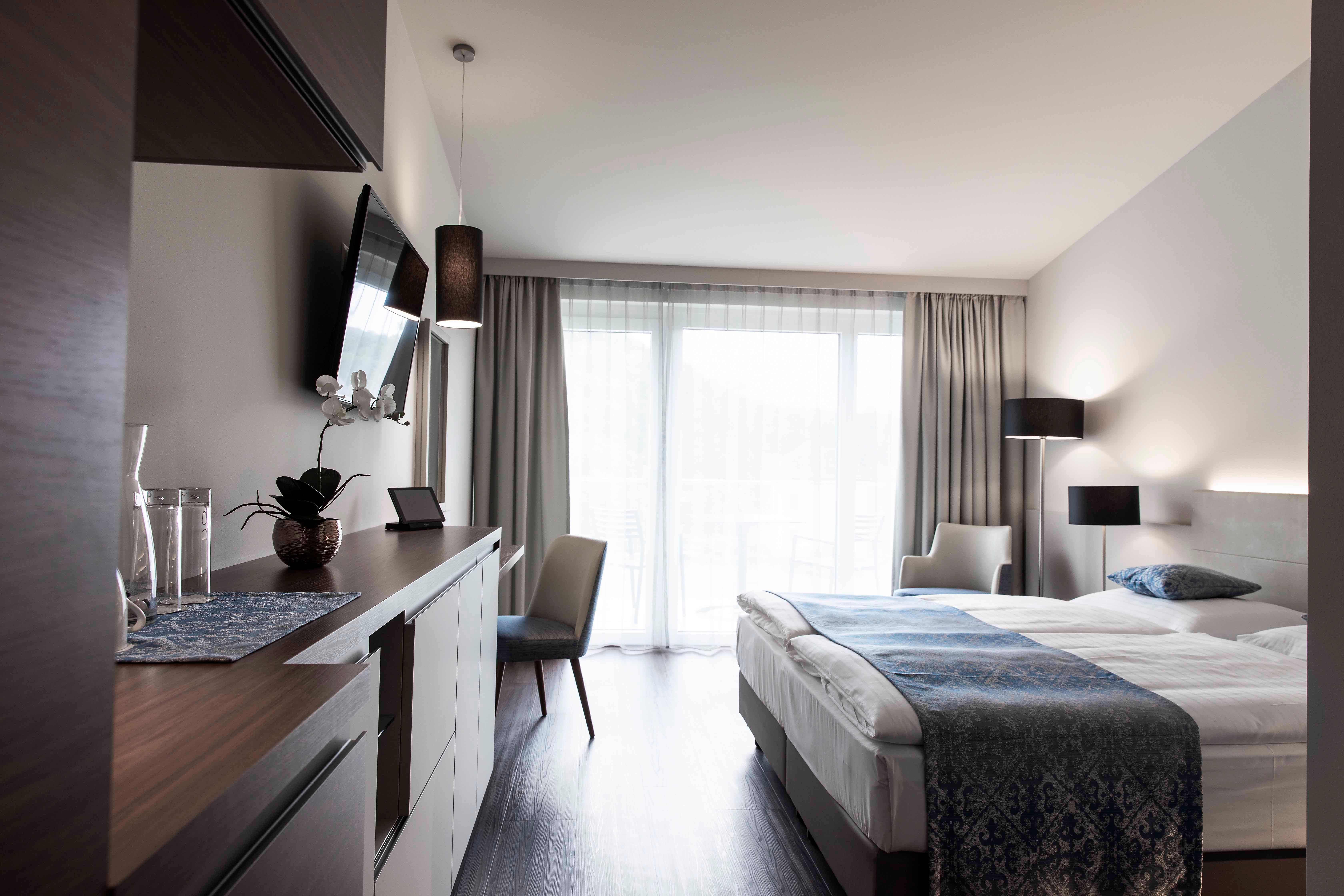 Modernes Hotelzimmer mit Doppelbett, Fernseher, Schreibtisch und großen Fenstern mit Vorhängen.