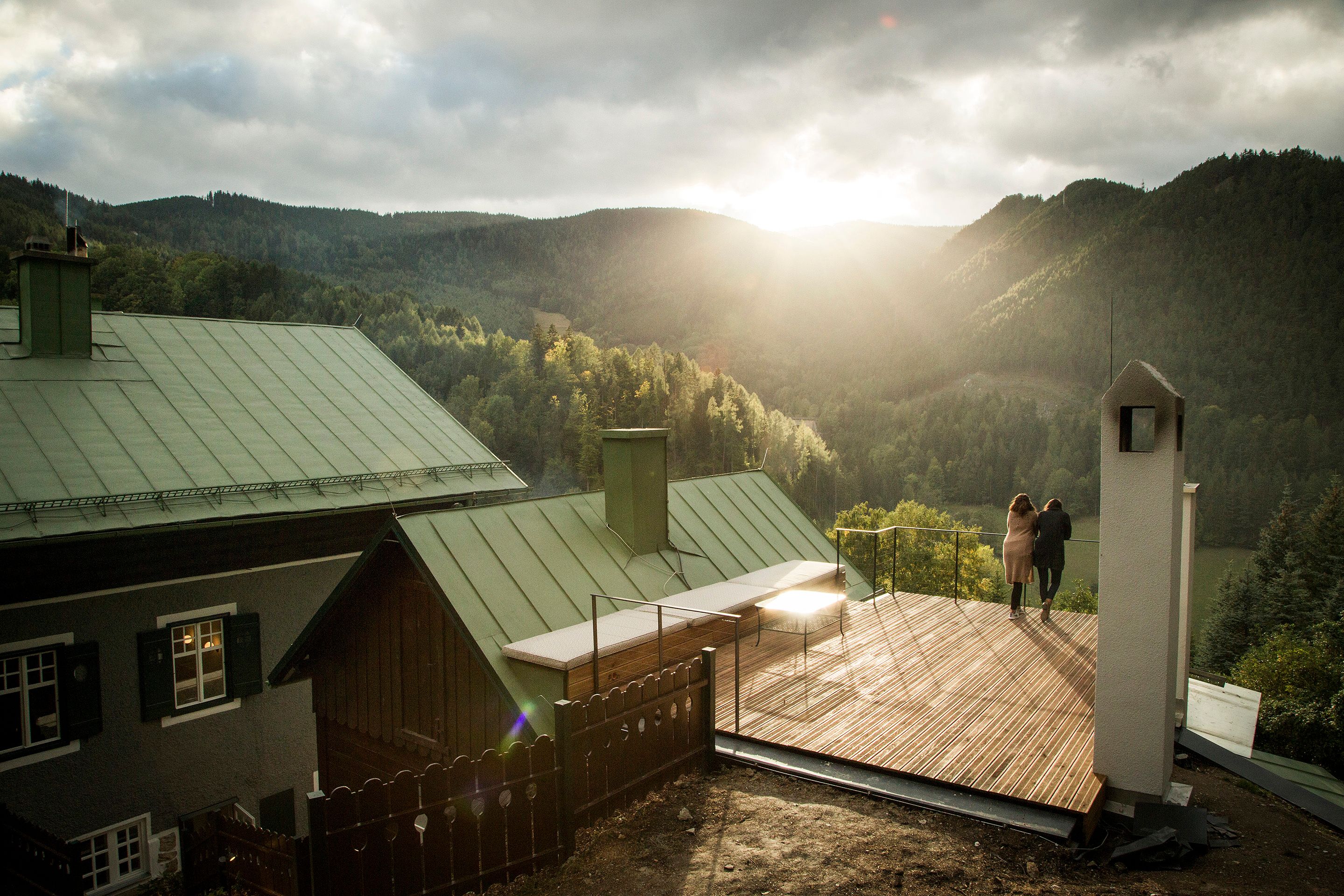 Zwei Personen stehen auf einer Terrasse mit Blick auf bewaldete Berge bei Sonnenuntergang.