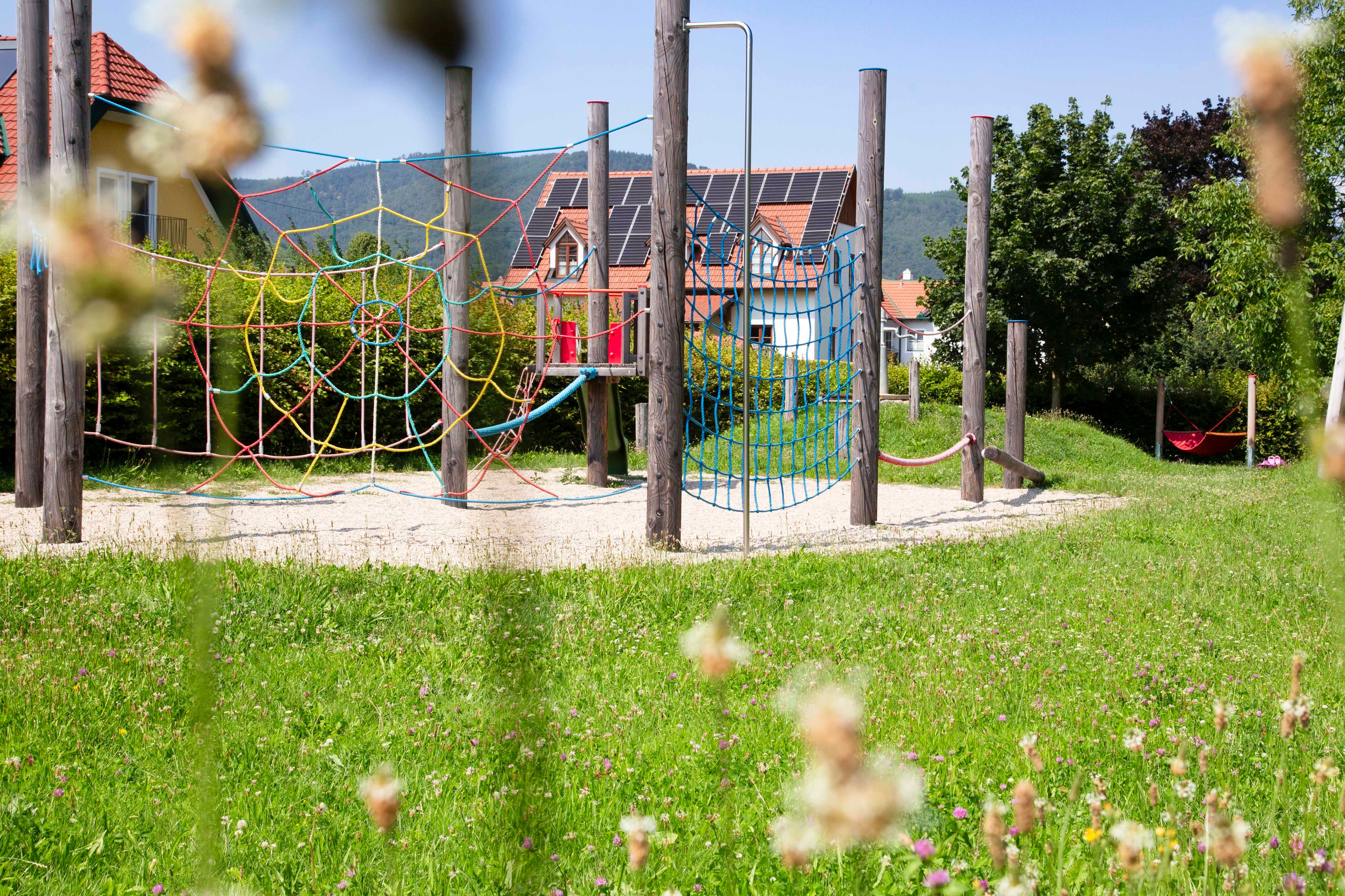 Ein Abenteuerspielplatz mit Kletternetzen und Holzpfosten, umgeben von grüner Wiese und Häusern im Hintergrund.