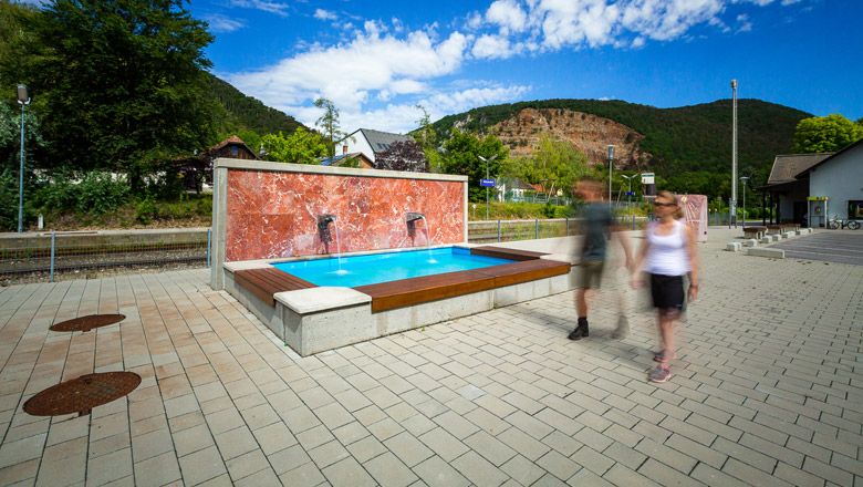 Moderner Brunnen auf einem Platz mit zwei unscharfen Personen im Vordergrund, umgeben von Bäumen und Hügeln.