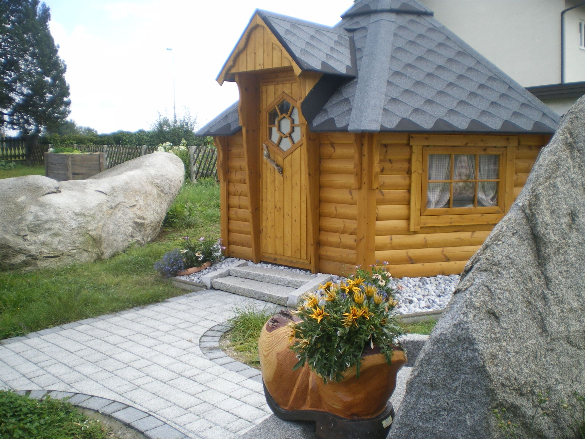 Eine kleine, hölzerne Grillhütte mit grauem Dach, umgeben von Steinen und Pflanzen.