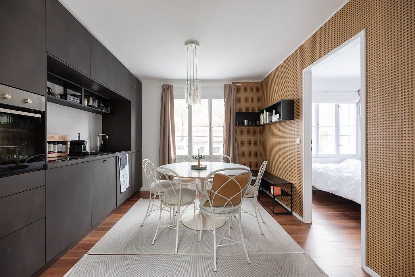 Moderne Küche mit dunklen Schränken, rundem Esstisch und weißen Stühlen, angrenzend an ein Schlafzimmer.