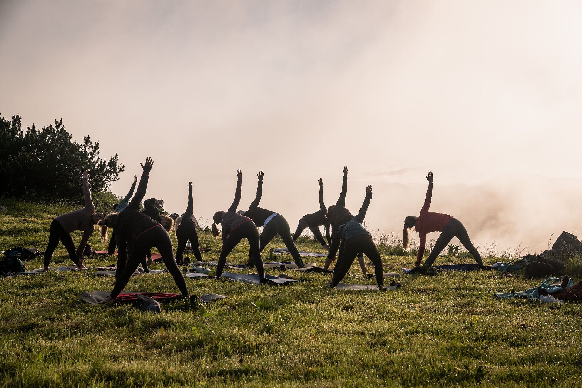 Inmitten der majestätischen Berge praktizieren Menschen Yoga, während die sanfte Morgenbrise durch die Wiesen weht. Die harmonischen Bewegungen der Teilnehmer verschmelzen mit der atemberaubenden Kulisse, die von Nebelschwaden umhüllt ist. Ein Ort der Ruhe und des inneren Friedens, ideal für eine Auszeit in der Natur.