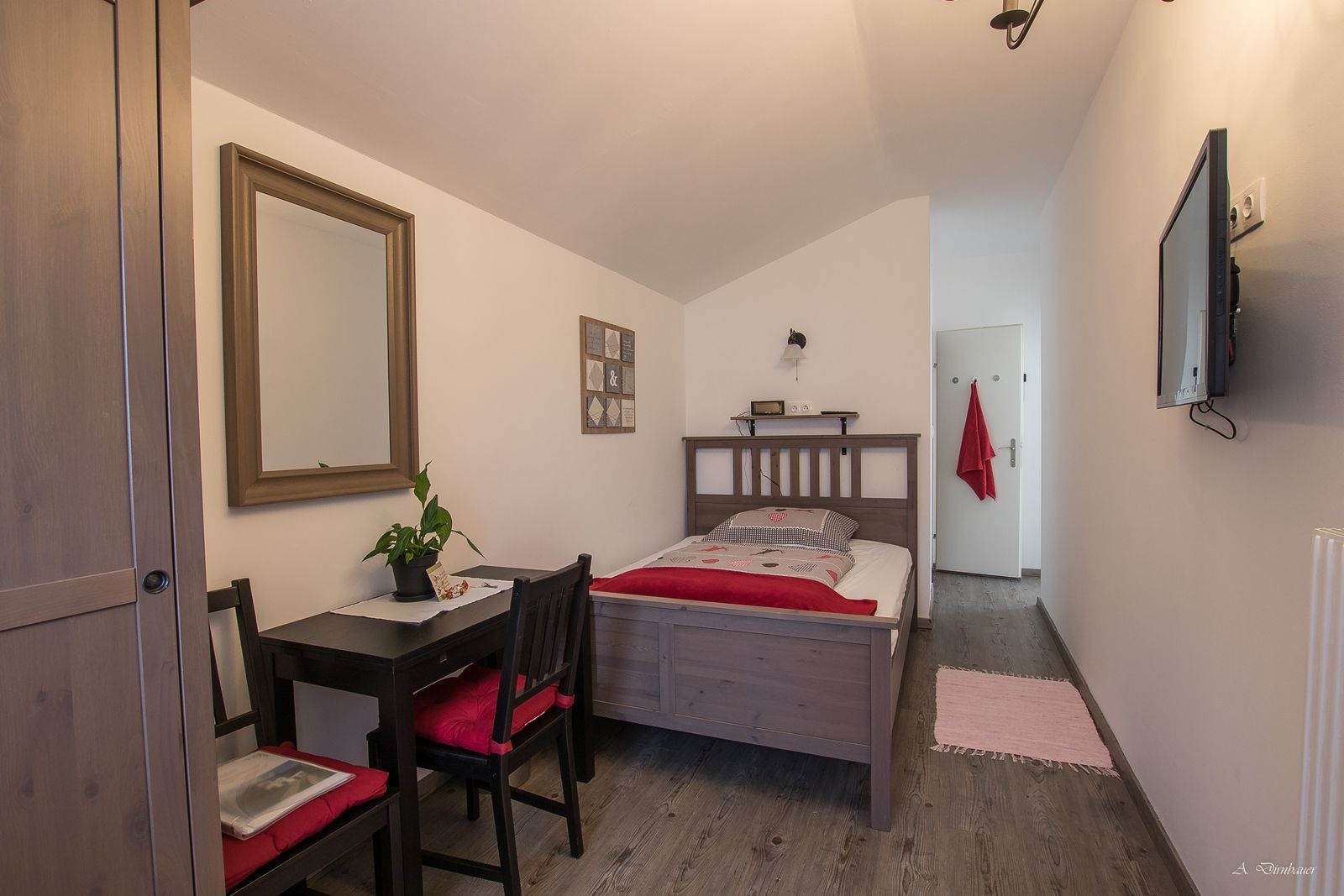 Ein gemütliches Einzelzimmer mit Holzmöbeln, einem Bett, einem kleinen Tisch mit Stühlen, einem Spiegel und einem Fernseher an der Wand.