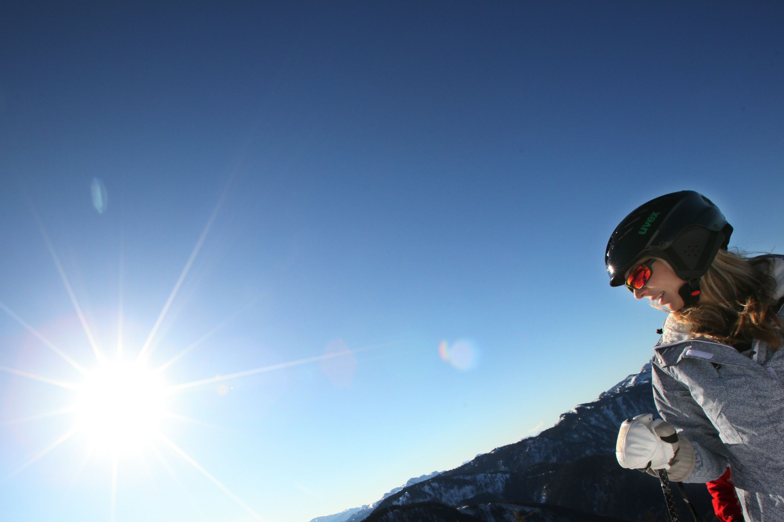 Eine Person mit Helm und Skibrille steht vor einem sonnigen, klaren Himmel in einer Berglandschaft.