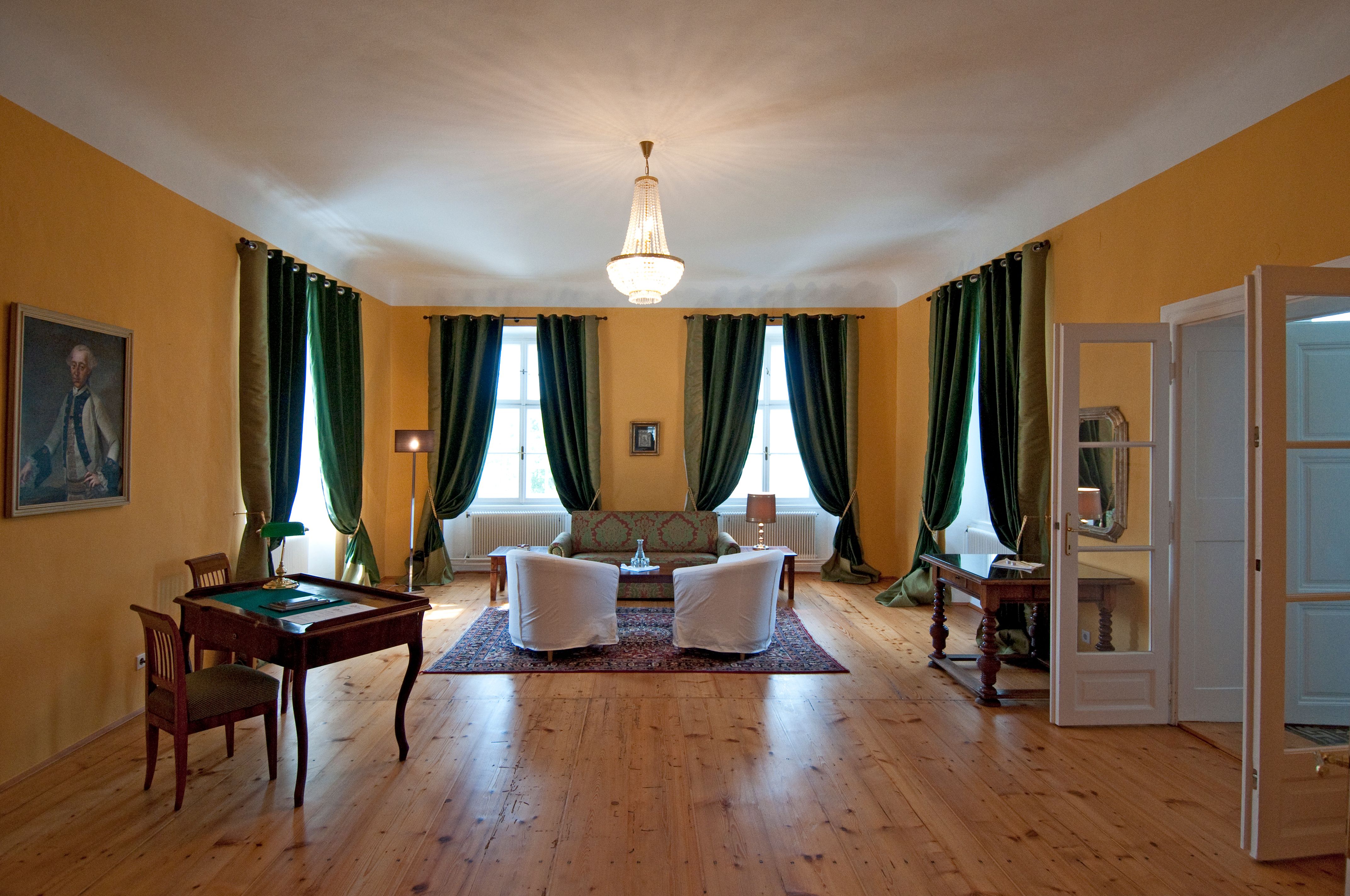 Elegantes Zimmer mit Holzboden, grünen Vorhängen, zwei Sesseln, einem Sofa und einem Schreibtisch. Ein Kronleuchter hängt von der Decke.