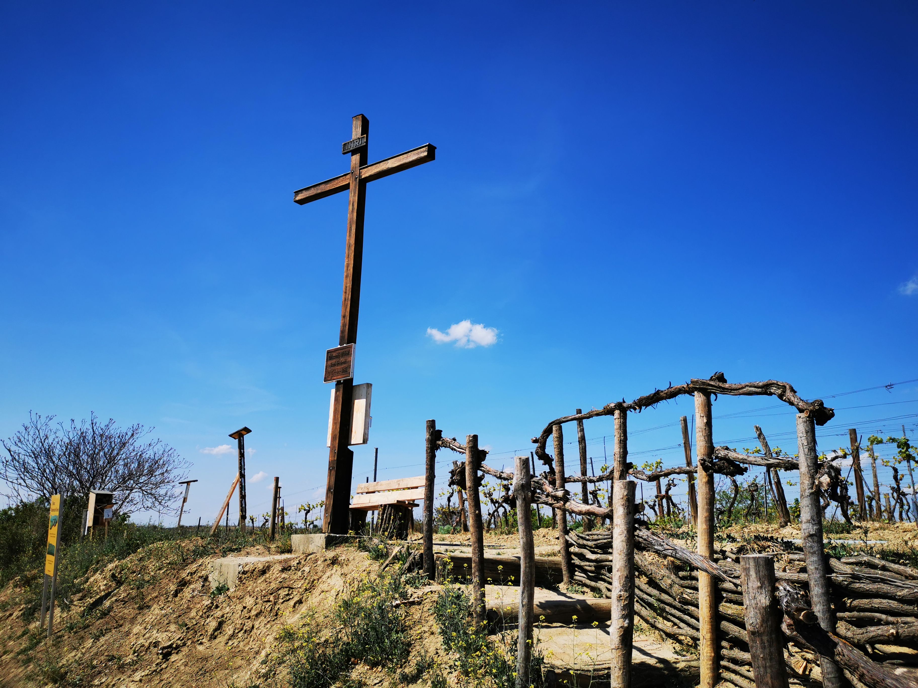 Holzkreuz auf einem Hügel mit Weinreben und blauem Himmel im Hintergrund.