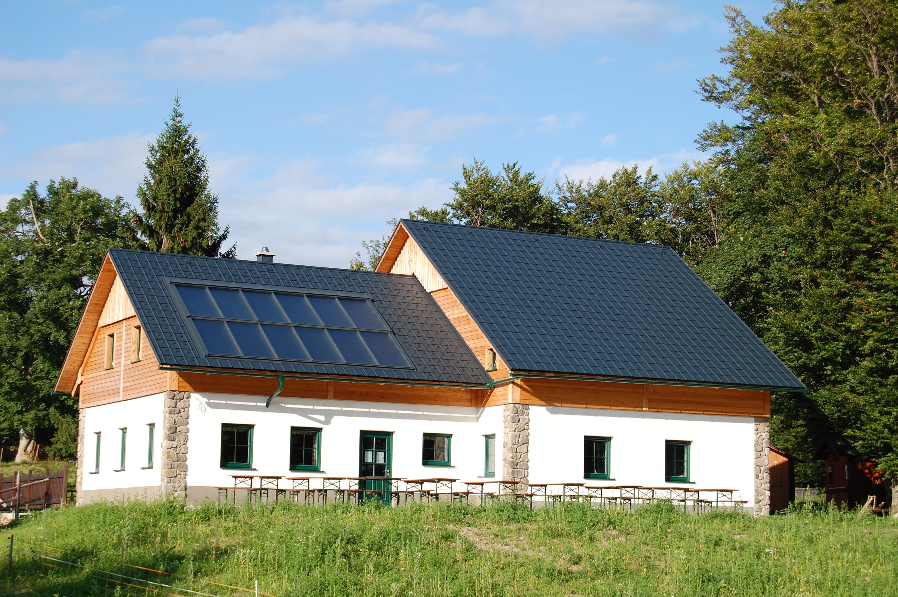 Eine Hütte mit Solarpanelen auf dem Dach, umgeben von Bäumen und Wiese.