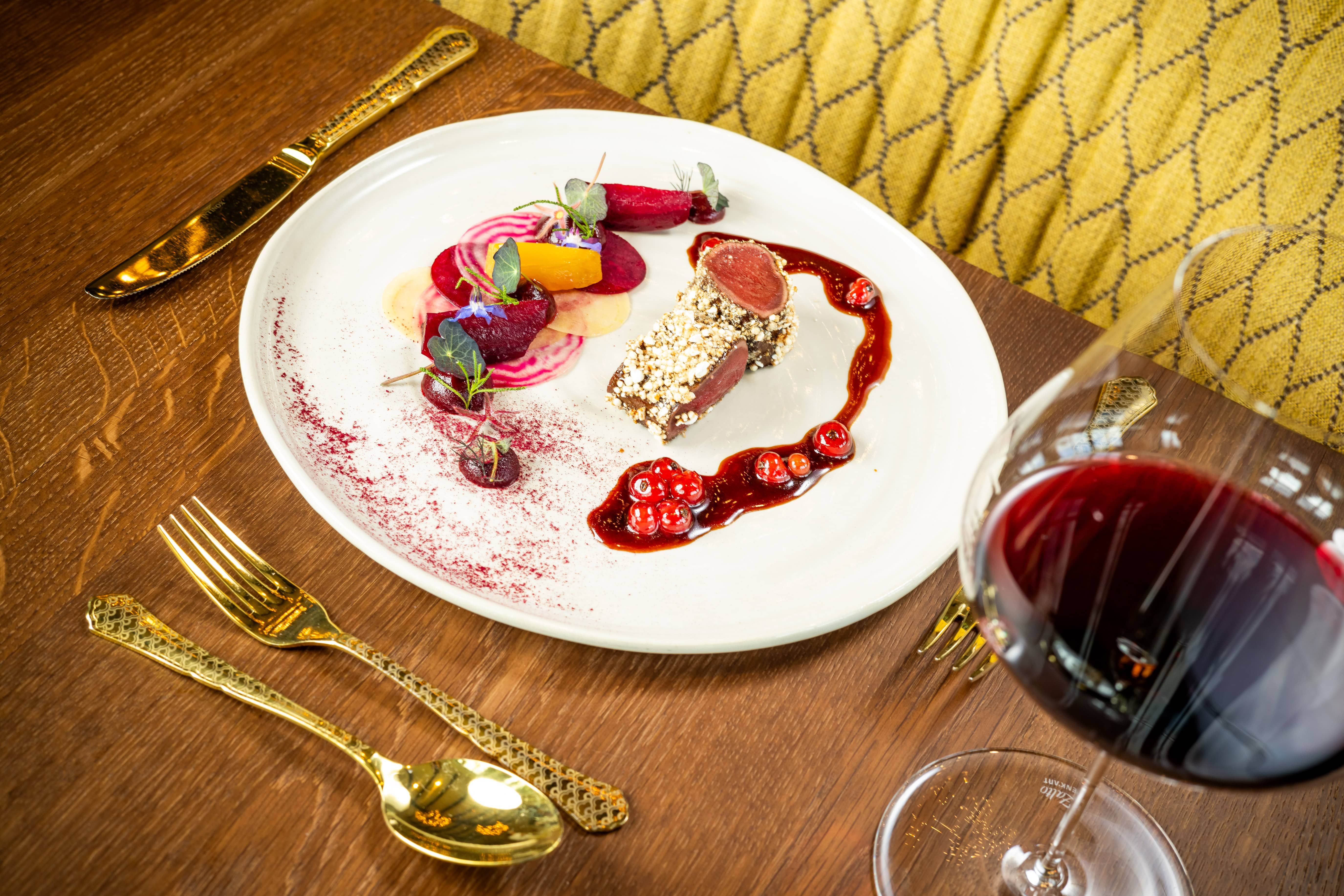 Ein Teller mit kunstvoll angerichtetem Rehrücken, Beilagen und einer Rotweinsoße, daneben ein Glas Rotwein auf einem Holztisch.