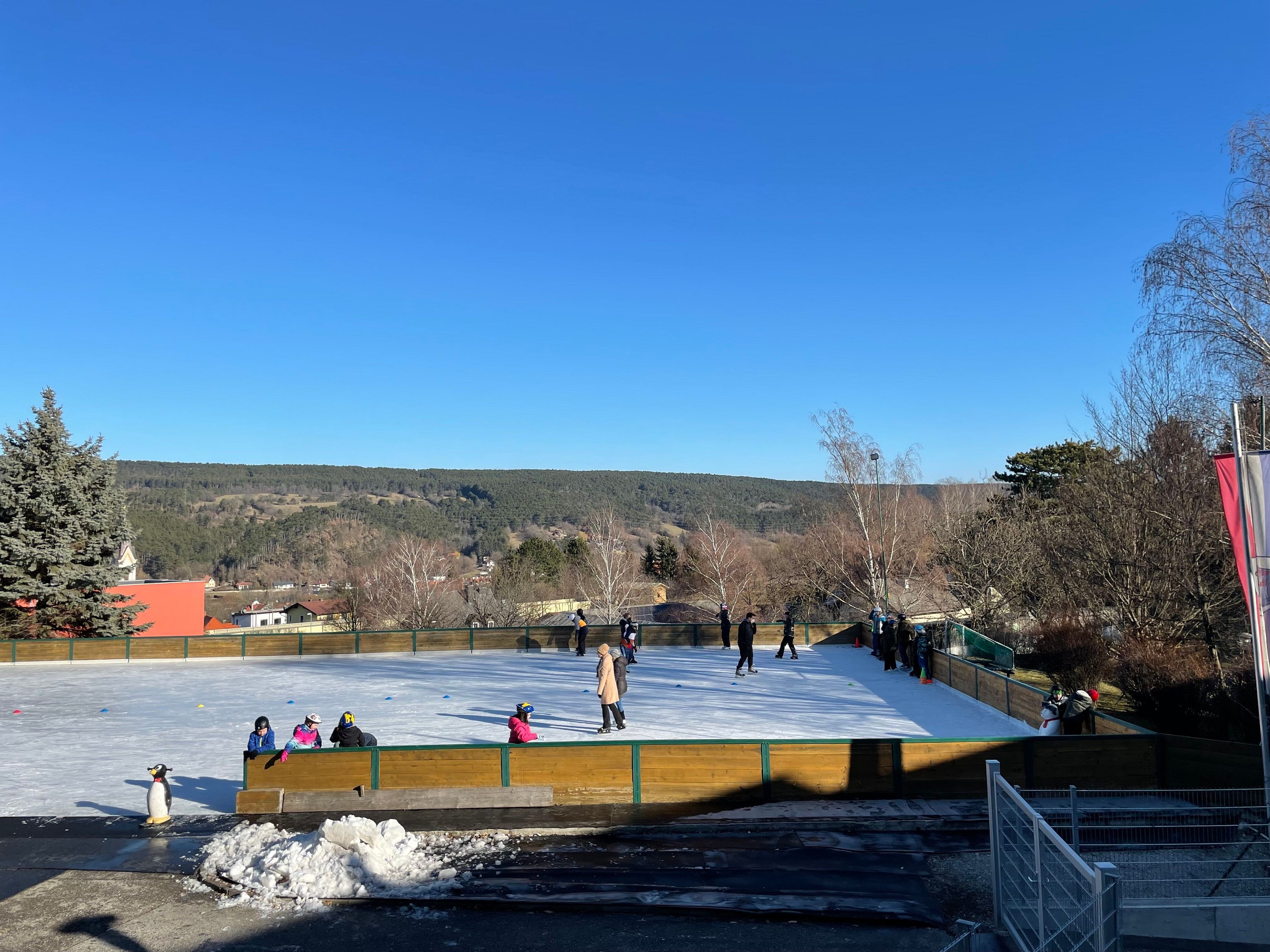 Eislaufplatz mit Menschen und Pinguinfigur, umgeben von Bäumen und Hügeln unter blauem Himmel.