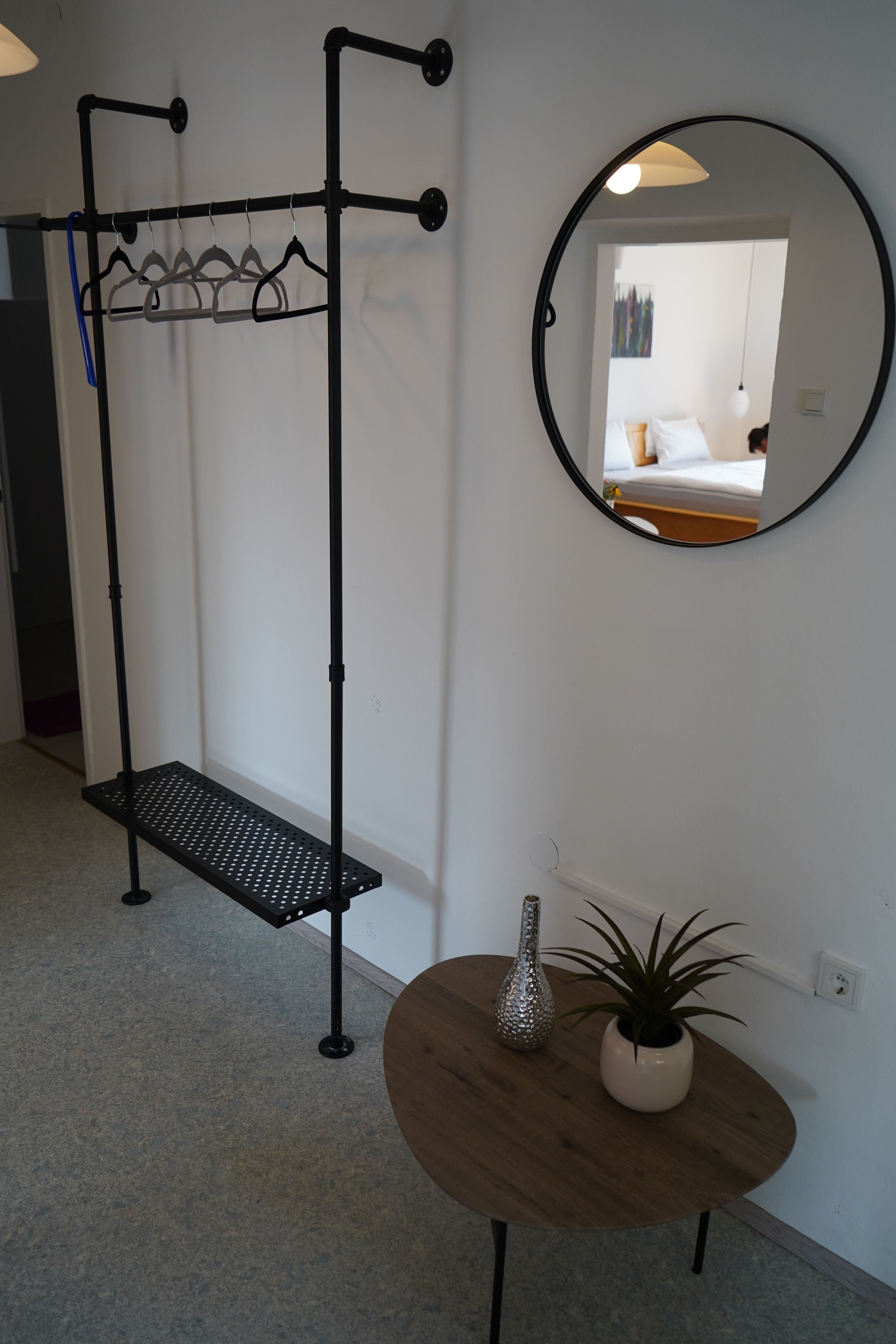 Innenansicht eines Zimmers mit Kleiderständer, rundem Spiegel und kleinem Tisch mit Pflanze und Vase.