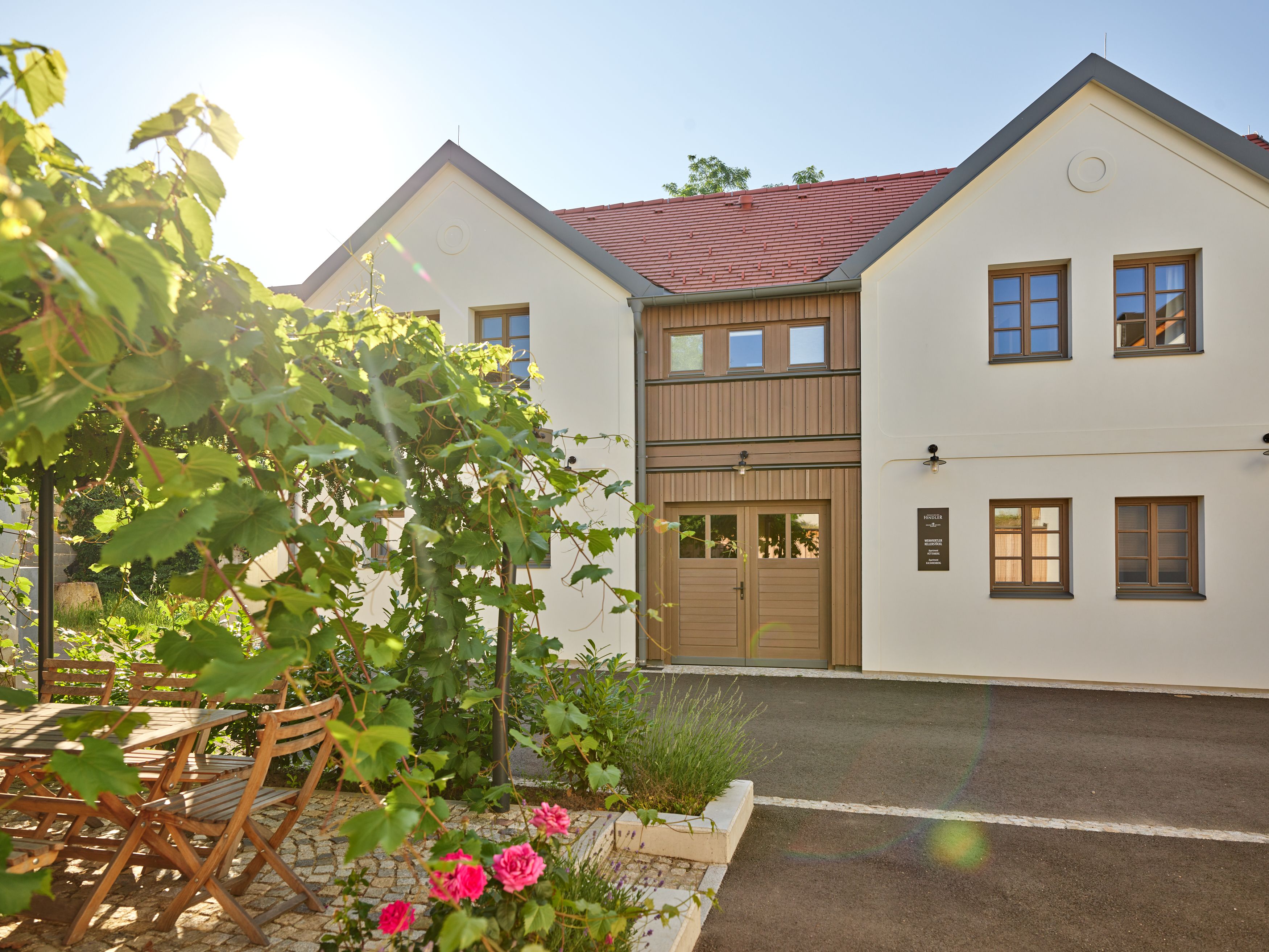 Ein modernes, zweistöckiges Gebäude mit weißen Wänden und roten Dachziegeln. Im Vordergrund ein Garten mit Holztisch, Stühlen und blühenden Pflanzen.