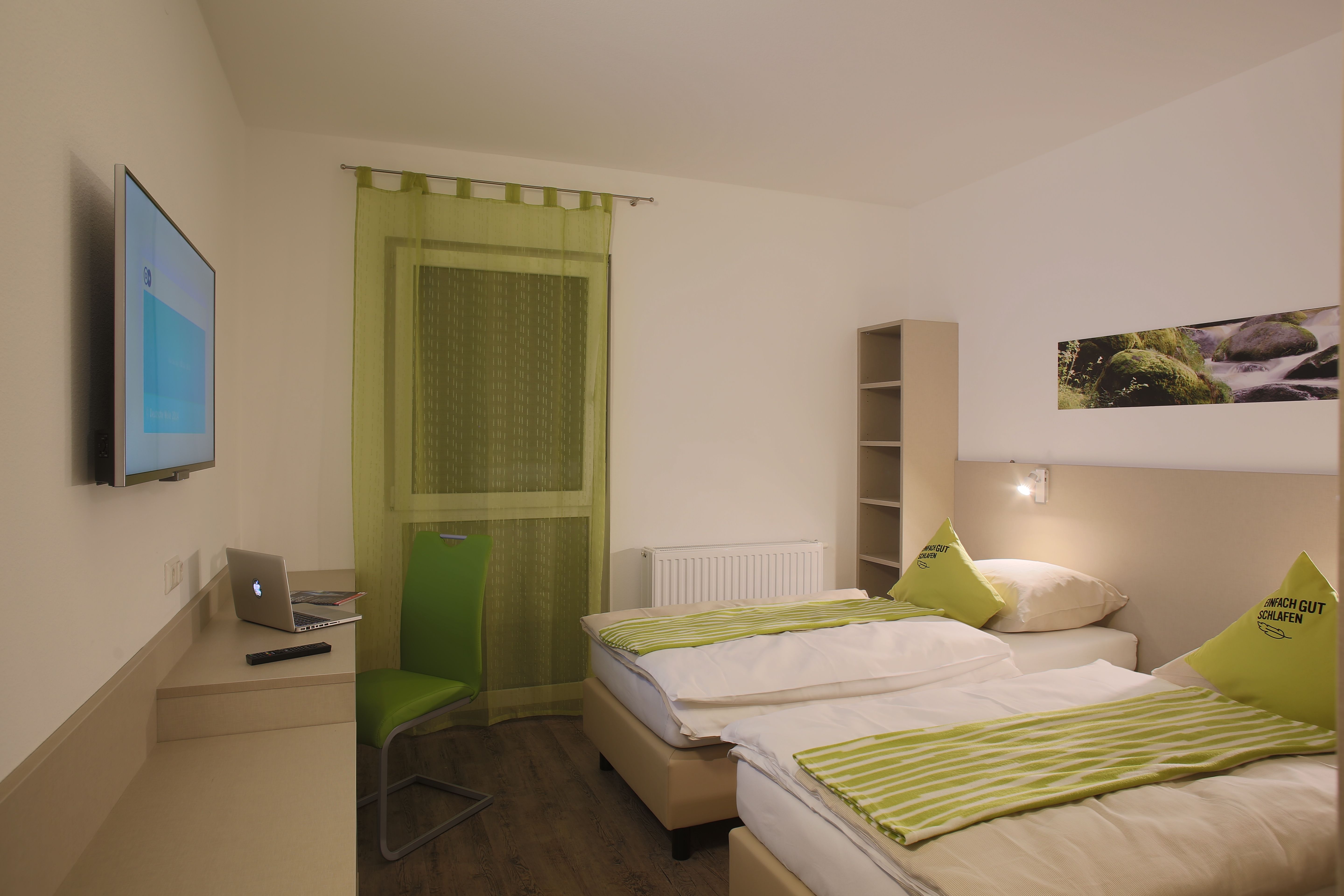 Modernes Motelzimmer mit zwei Betten, grünem Stuhl, Laptop auf Tisch und Wand-TV.